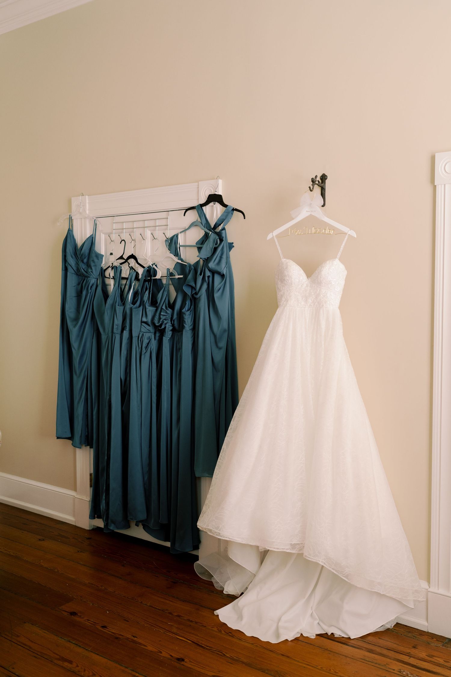 White wedding dress and teal bridesmaid dresses hanging on display against a beige wall.
