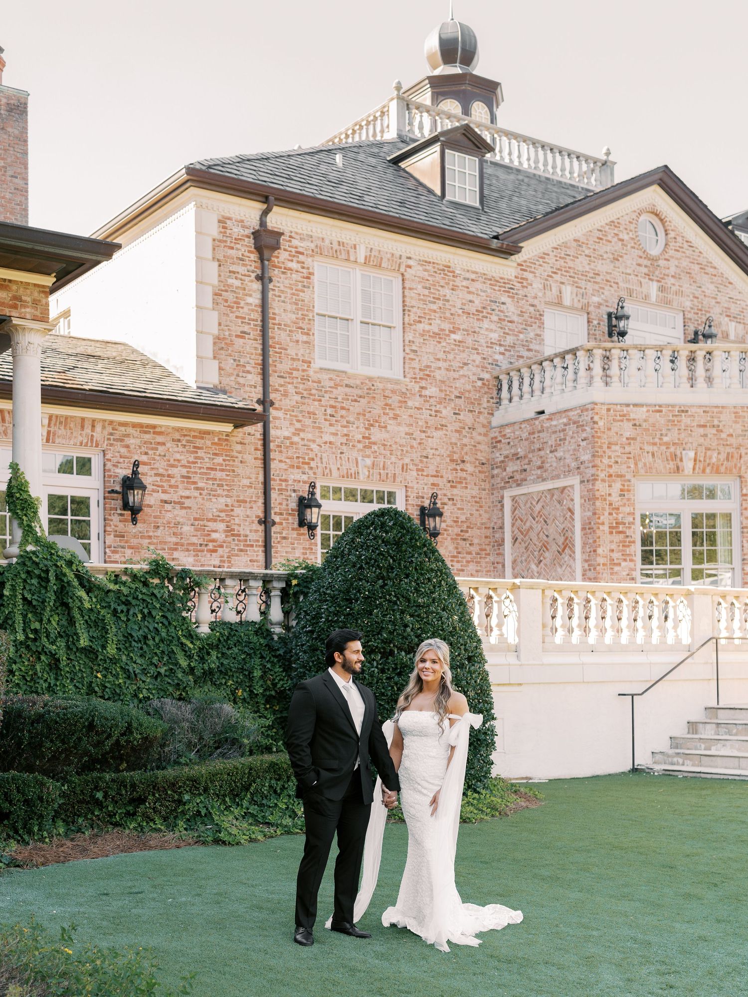 A couple poses on the lawn of an elegant brick mansion with white balustrades.
