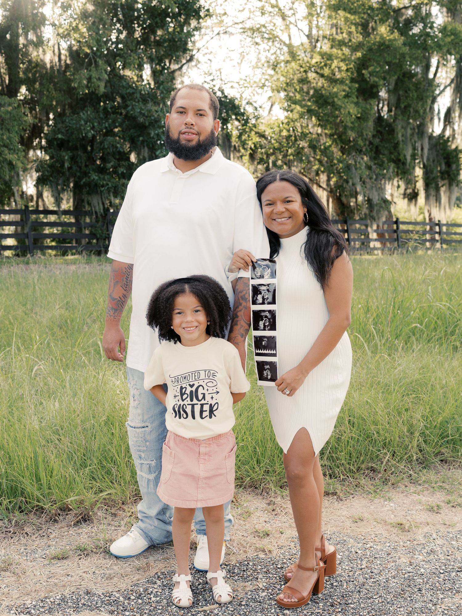A family displays pregnancy announcement signs on a rural path with trees in background.