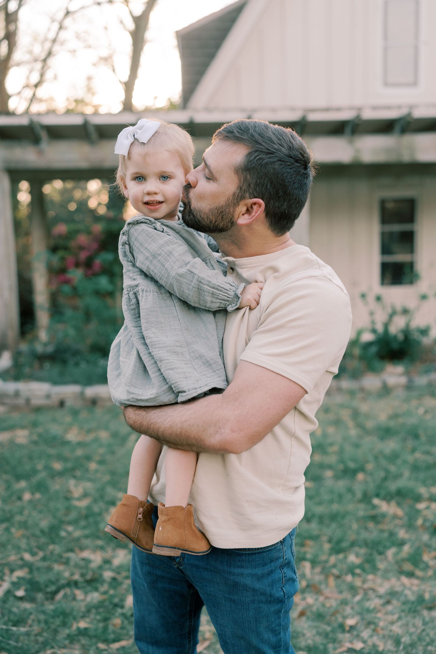 A parent holds and kisses a small child wearing gray clothing against a white building backdrop.