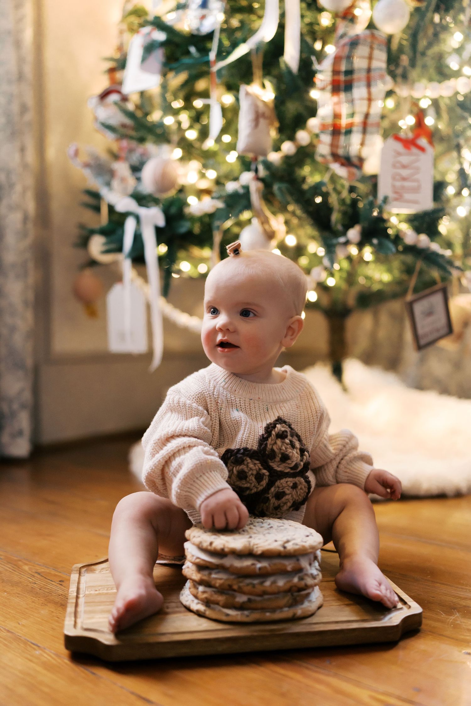 Baby in cream sweater sits by stacked pancakes near a decorated Christmas tree.