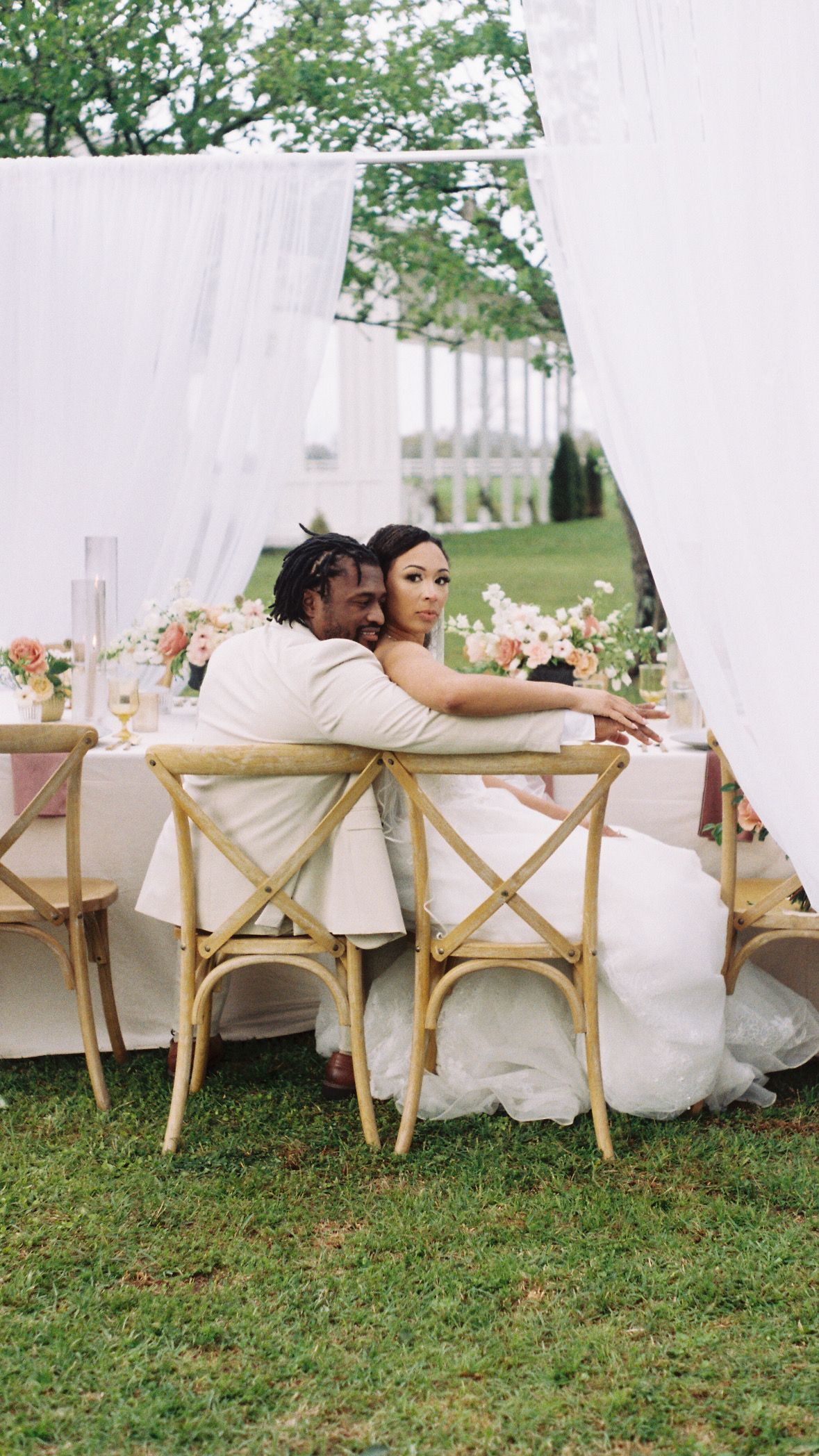Couple sitting at elegant outdoor wedding reception table with white draping and floral decorations.