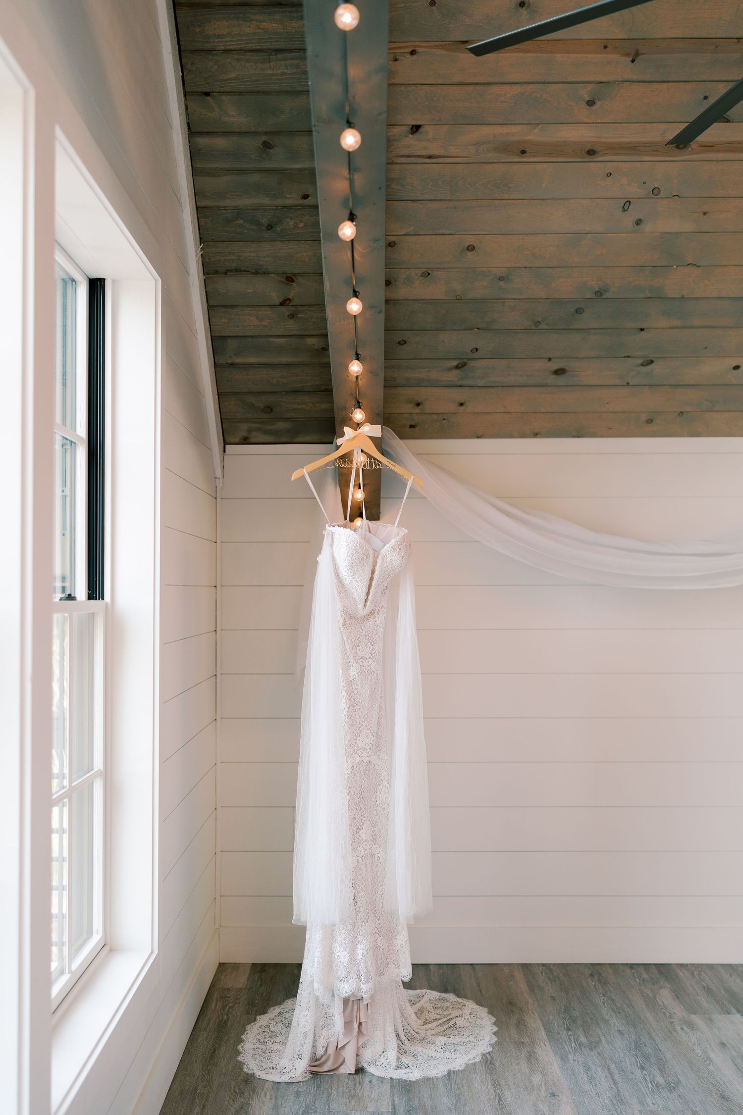 White lace wedding dress hangs elegantly against rustic wooden walls with string lights.