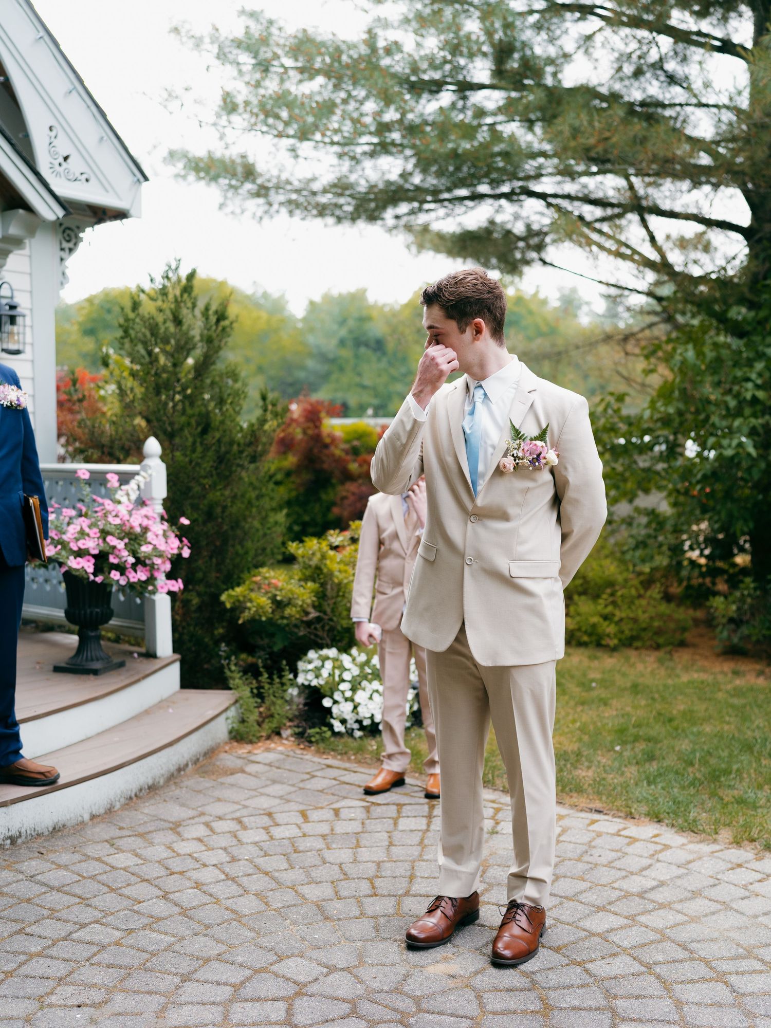 Wedding party members in light beige suits standing on stone patio near chapel entrance.