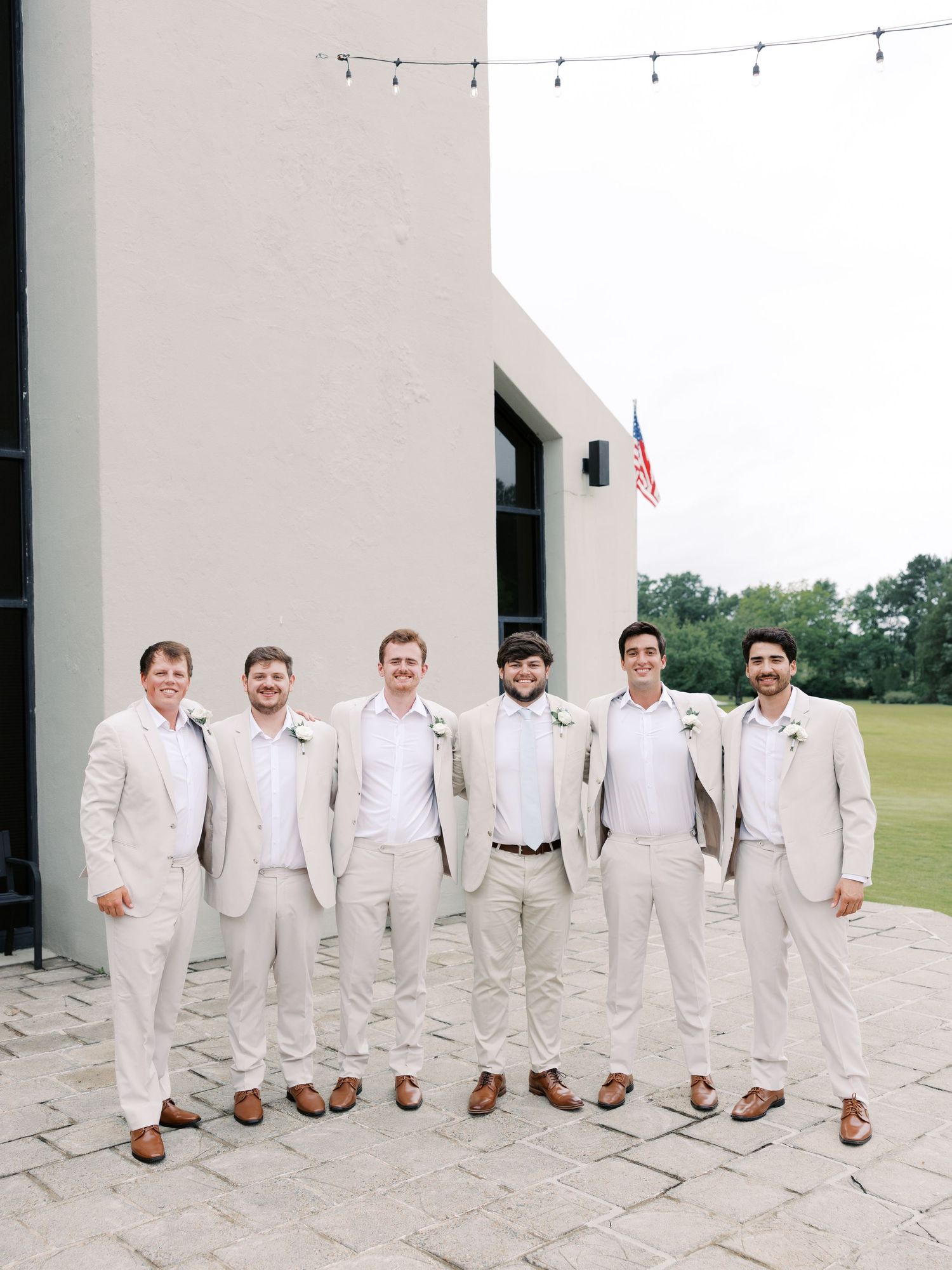 Wedding party members in light colored suits stand in a line outdoors.