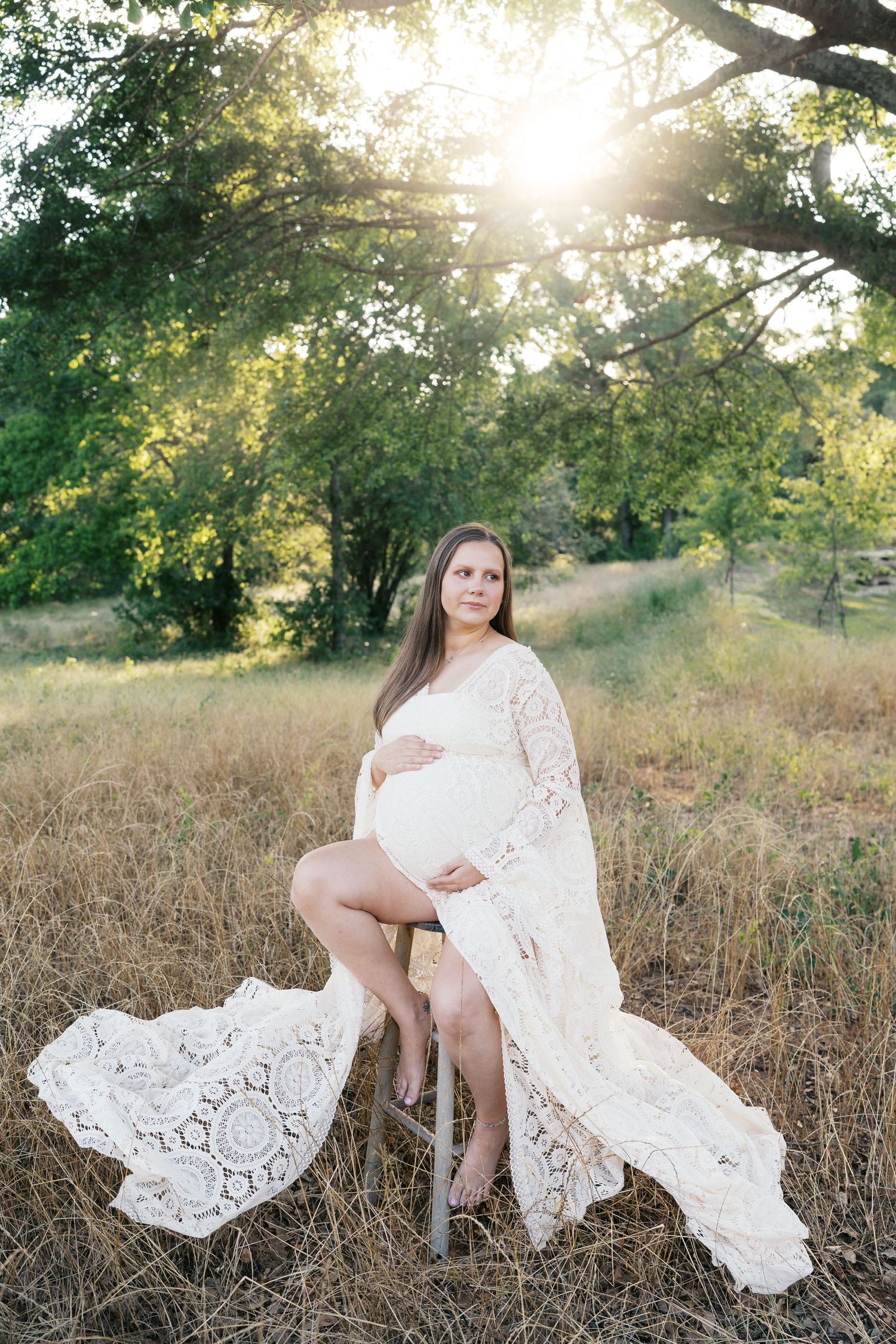 Ethereal maternity photoshoot in a field at golden hour featuring flowing white lace dress against natural scenery.