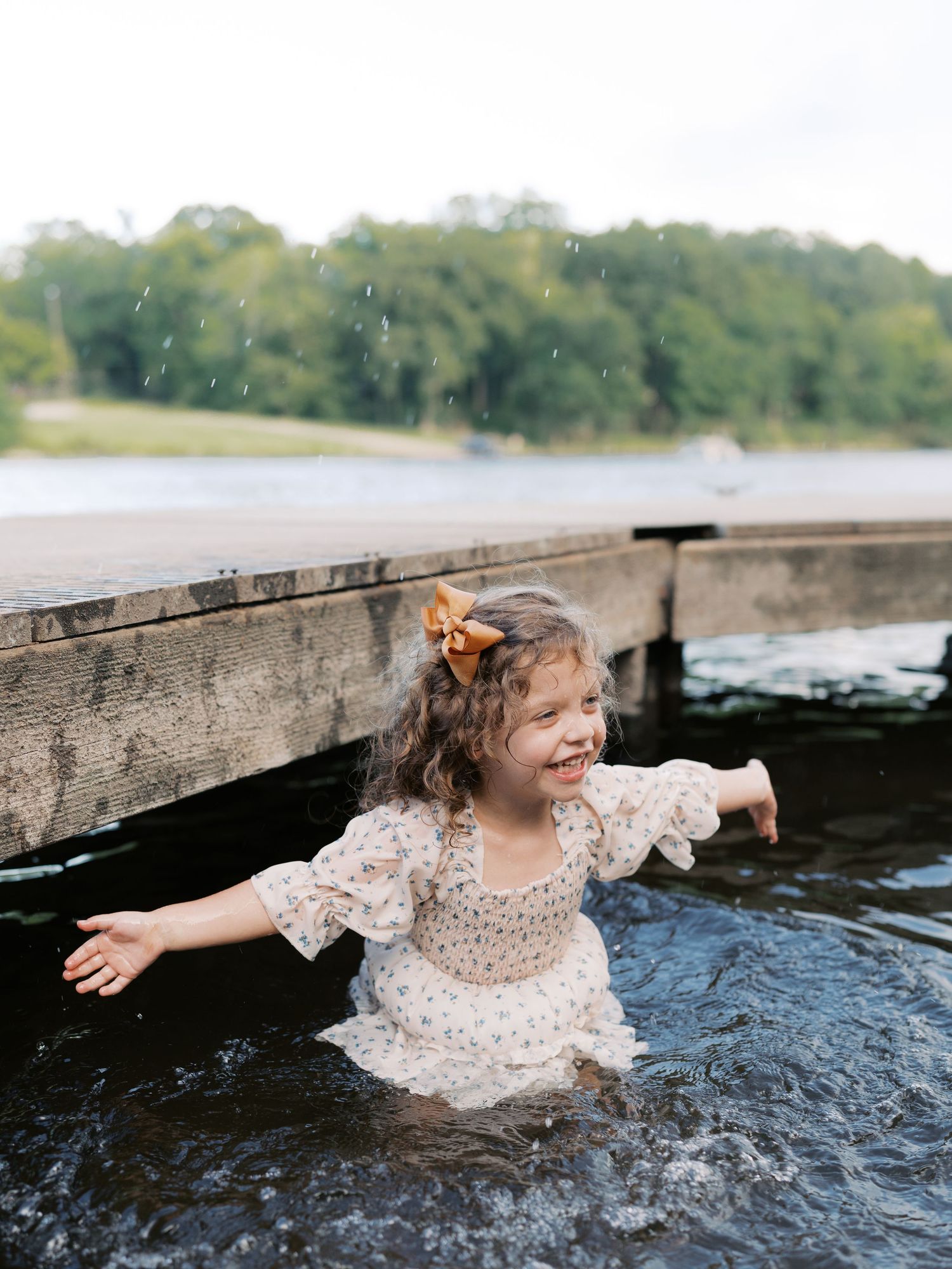 A young person in a light-colored dress splashes playfully in lake water near a wooden dock.