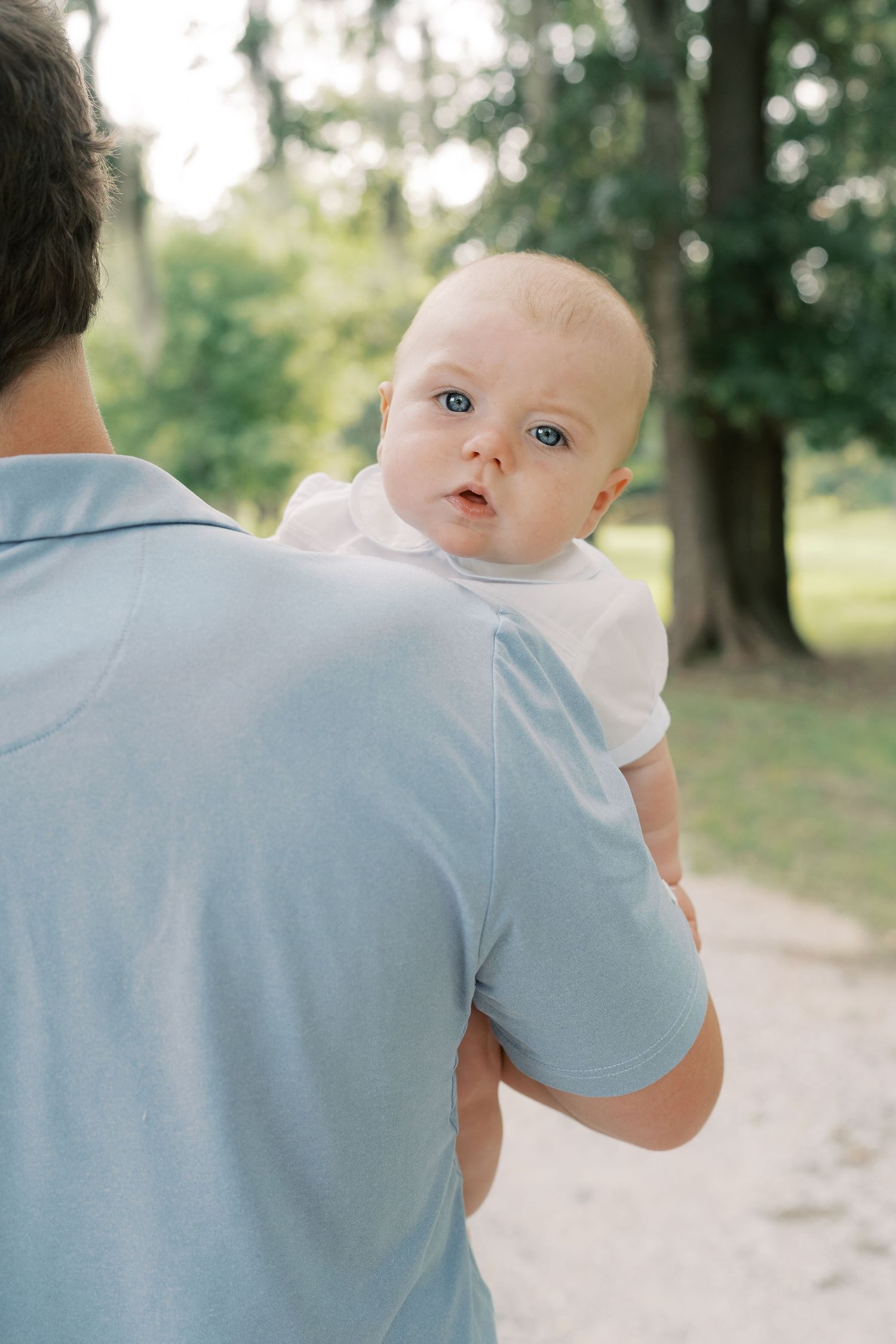 Baby in white clothing peeks over parent's shoulder on a wooded path.