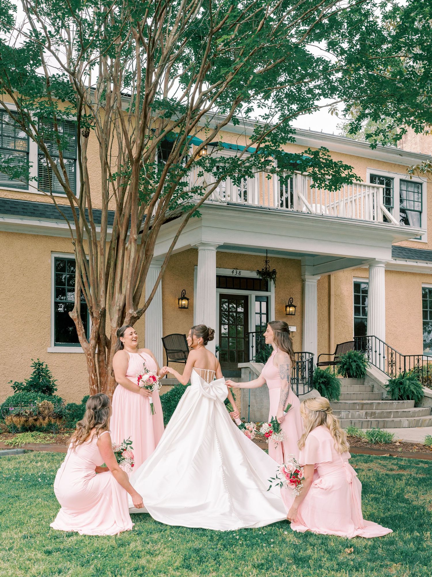 Bridesmaids in pink dresses gather around a bride in white on a mansion lawn.