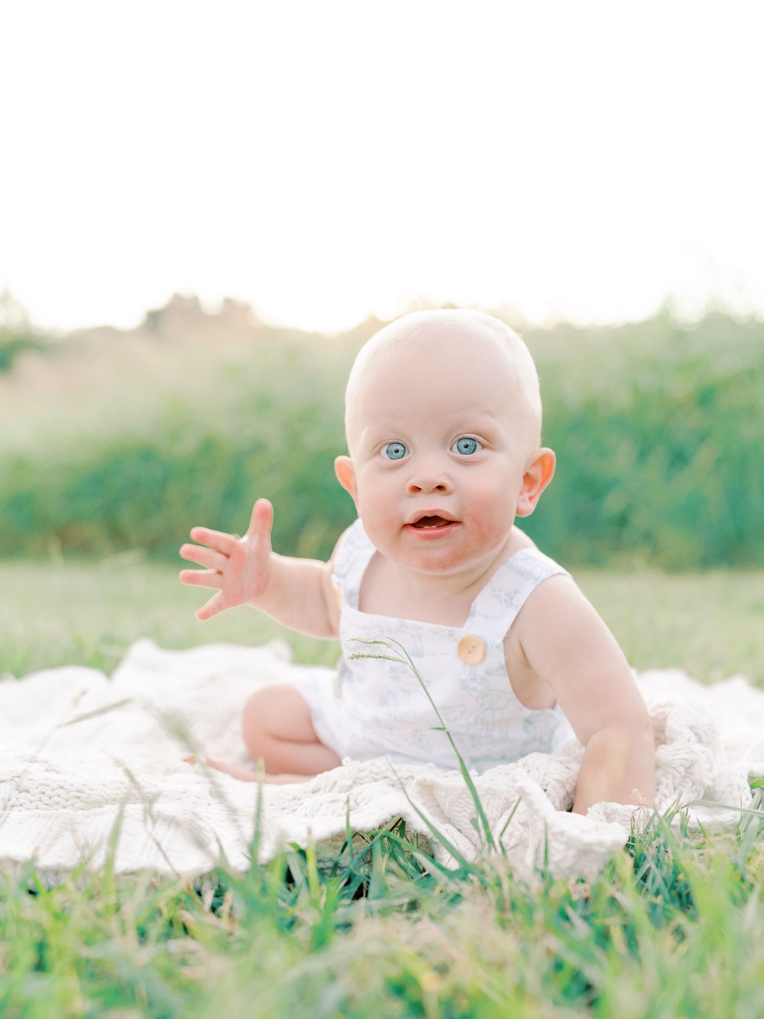 Baby with bright blue eyes lying on white blanket in grass during outdoor photo session.