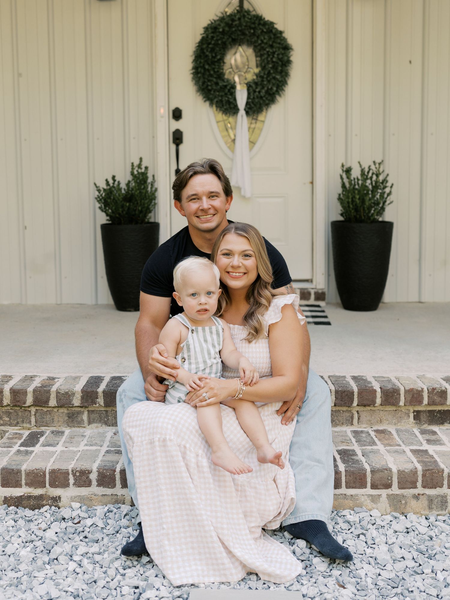Family sits together on brick steps of white house with decorative wreaths.