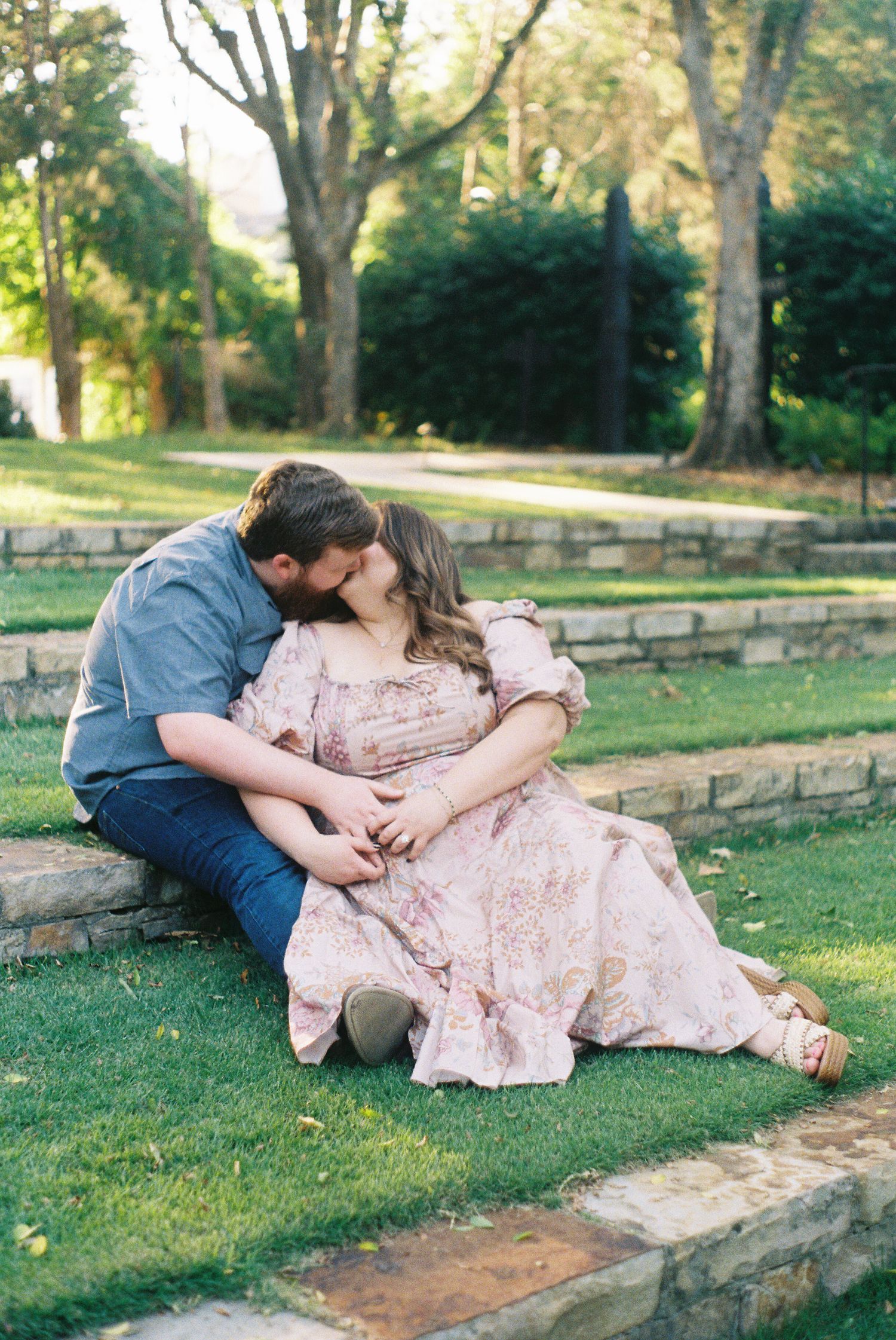 A couple in romantic poses lying on grass near a stone wall in a park setting during golden hour.
