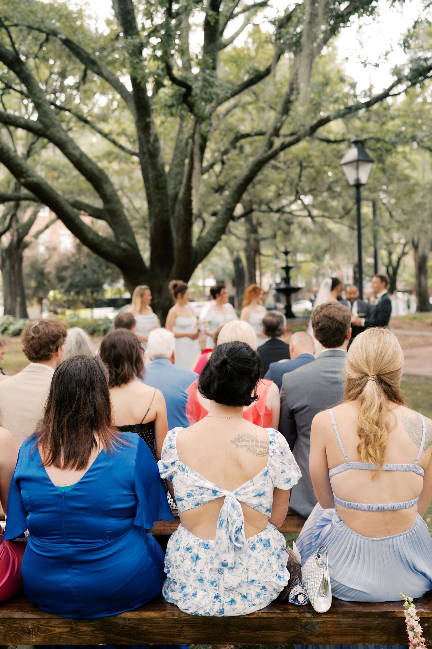 Bridesmaids in blue dresses sit together on a bench under large oak trees.