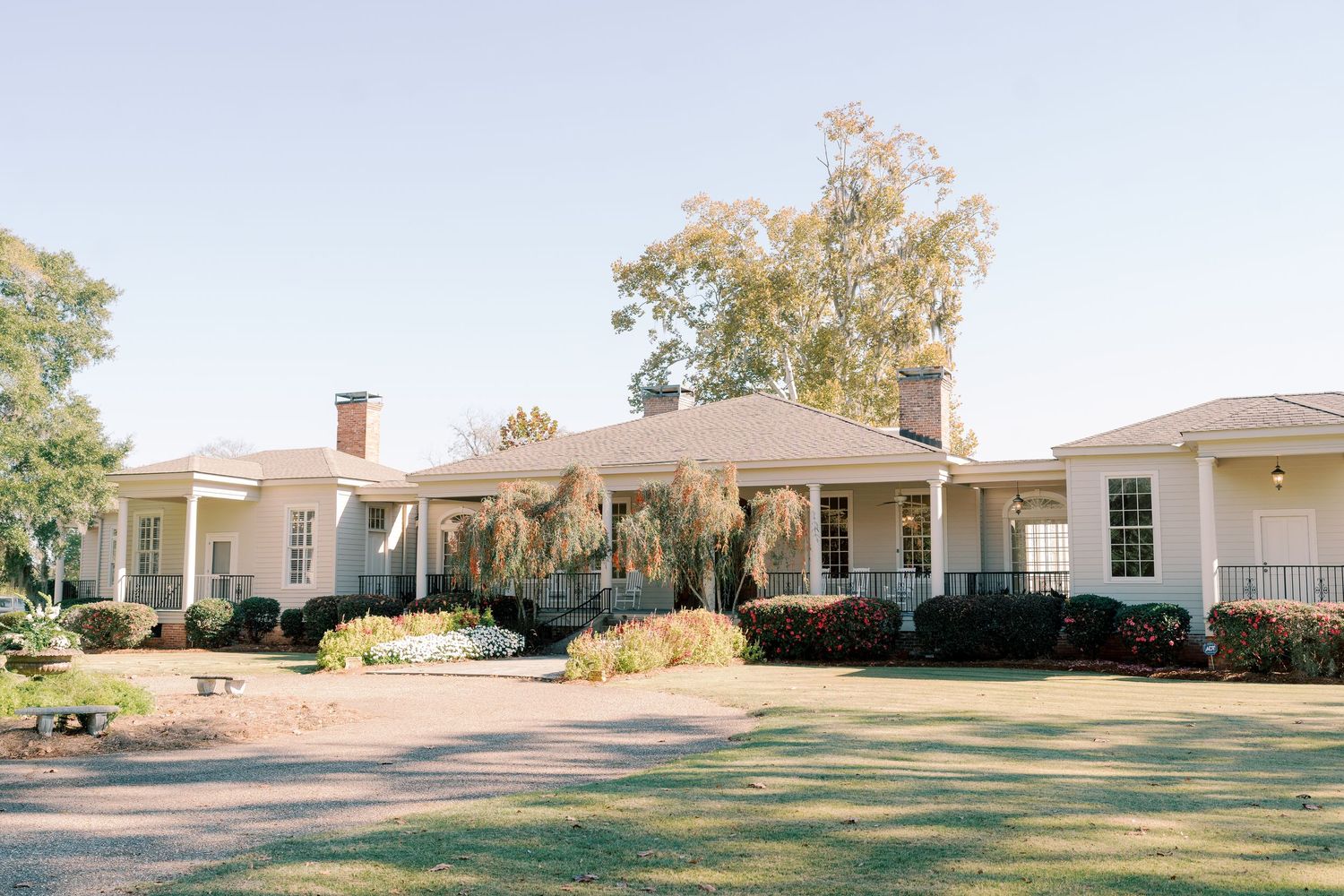 Elegant white southern-style mansion with wrap-around porch and manicured lawn on a sunny day.
