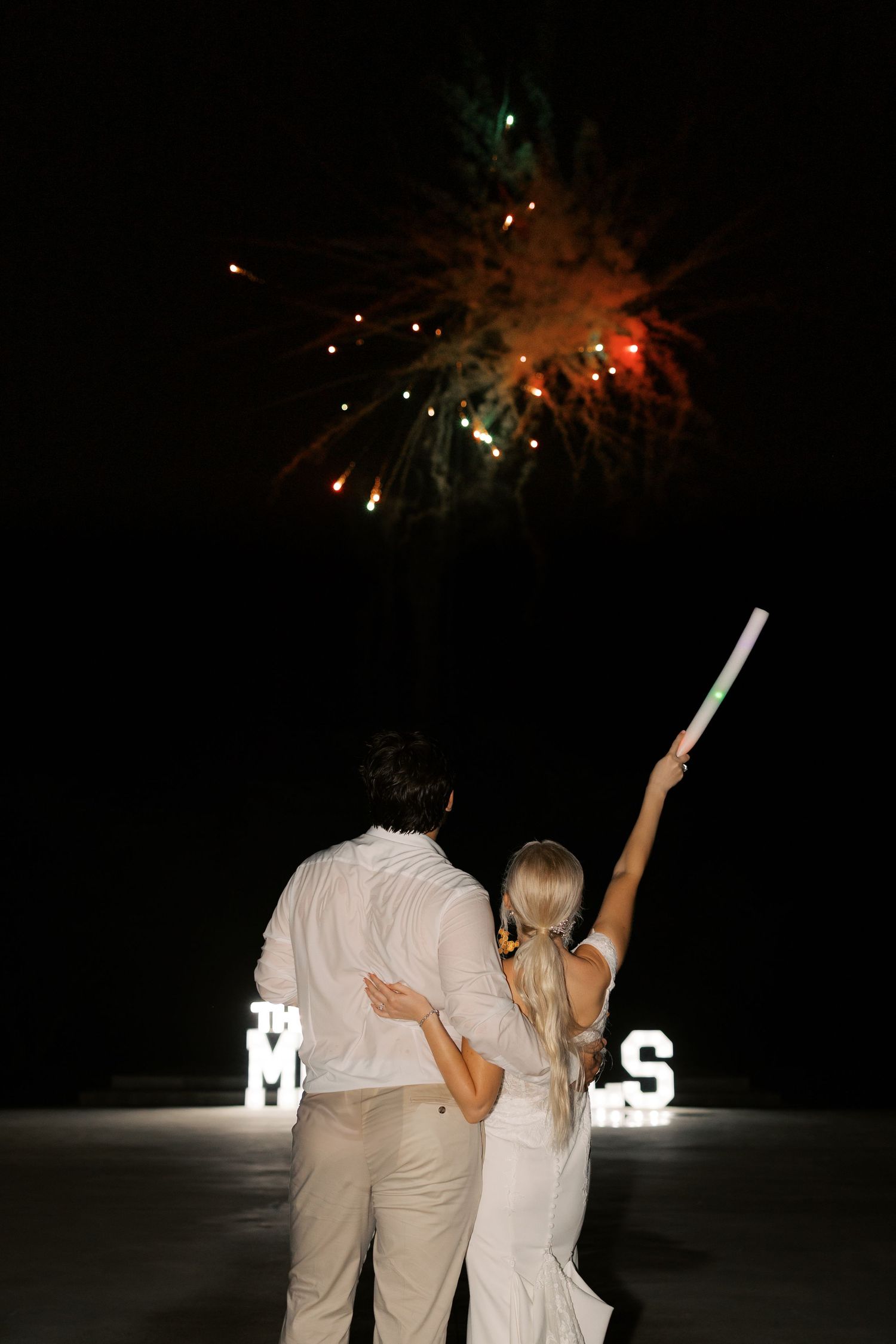 Wedding guests watch fireworks display at night with illuminated letters in the background.