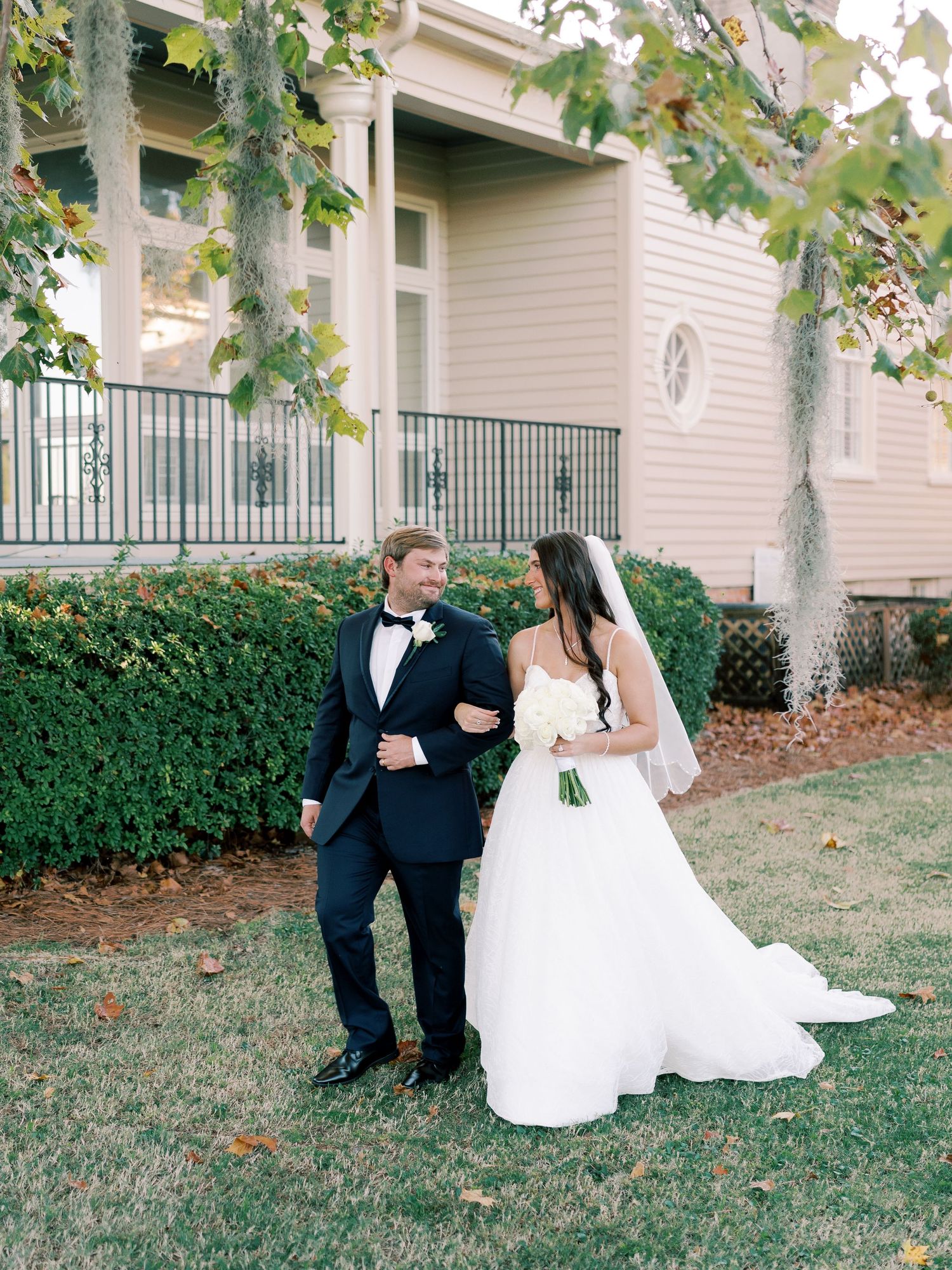 Bride in flowing white gown and groom in tuxedo share moment on manicured lawn.