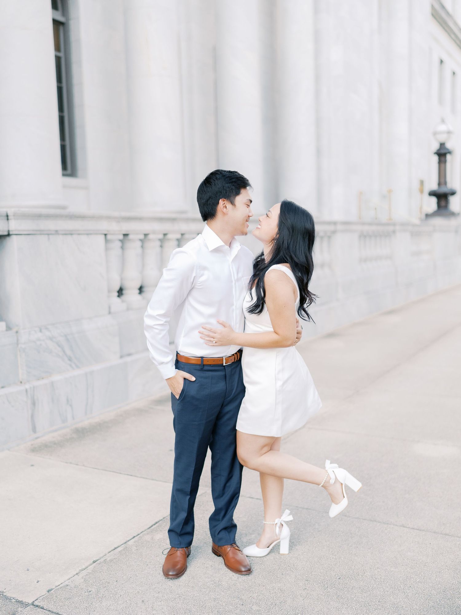 A couple in white outfits poses romantically against a marble building exterior.