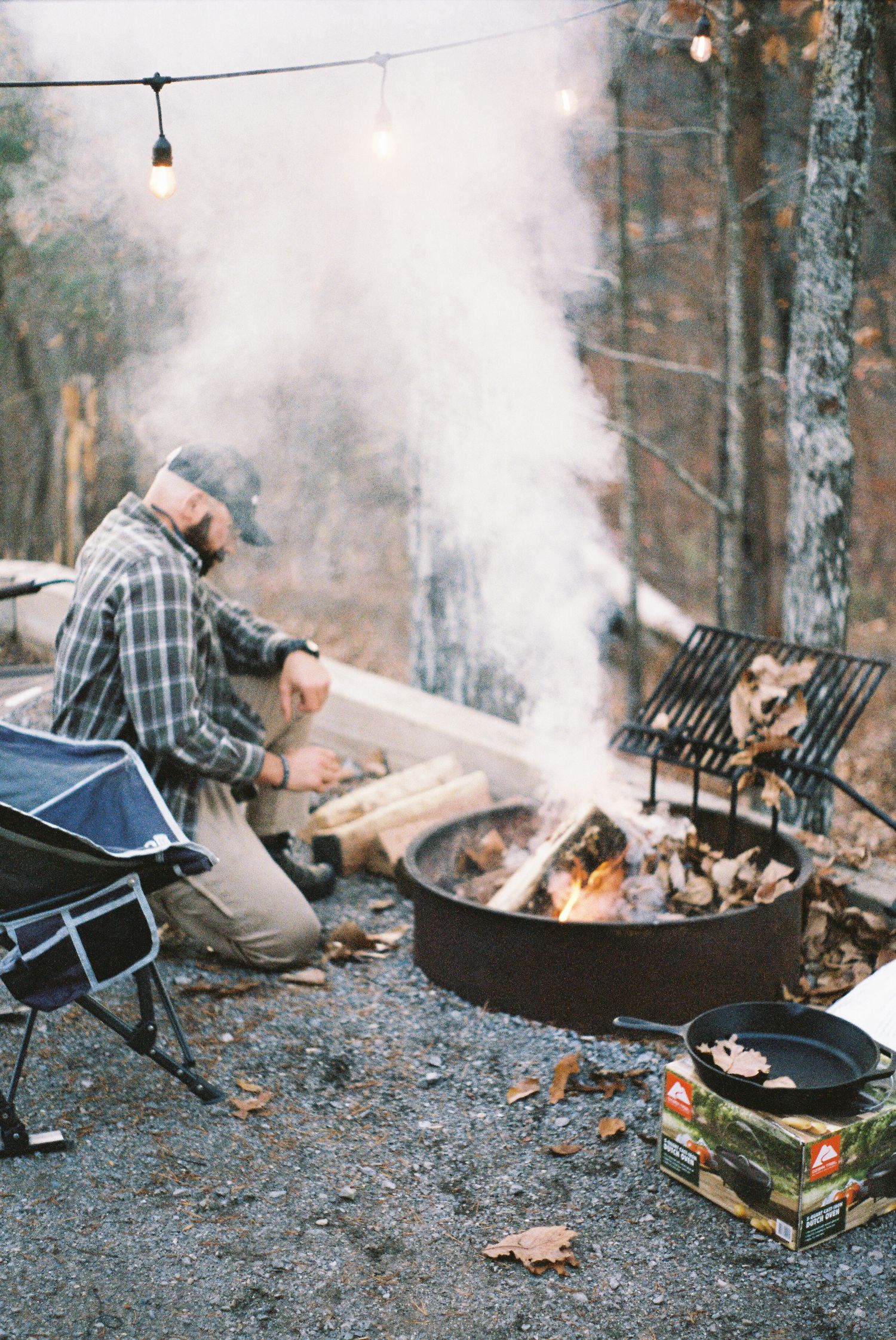 Campfire scene with smoke rising from a fire pit and camping chairs in a snowy forest setting.