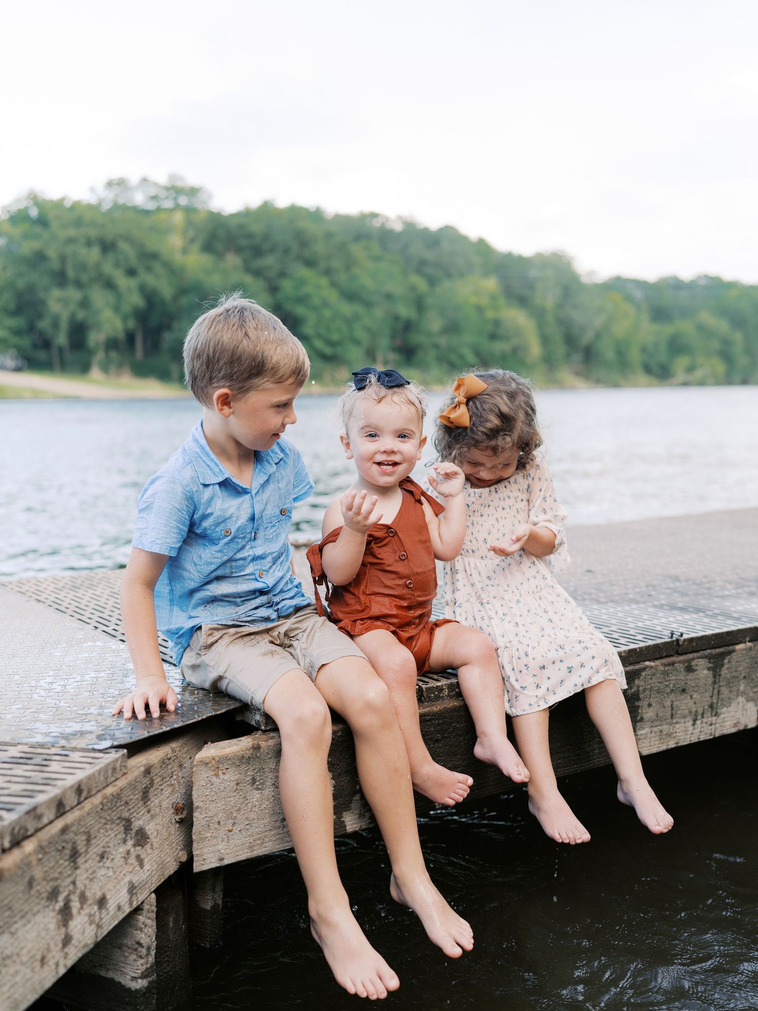 Three children sit together dangling their feet off a wooden dock over water.