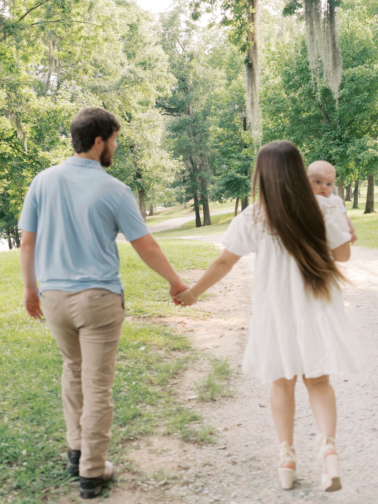 A couple holds hands while walking with their baby on a wooded trail.