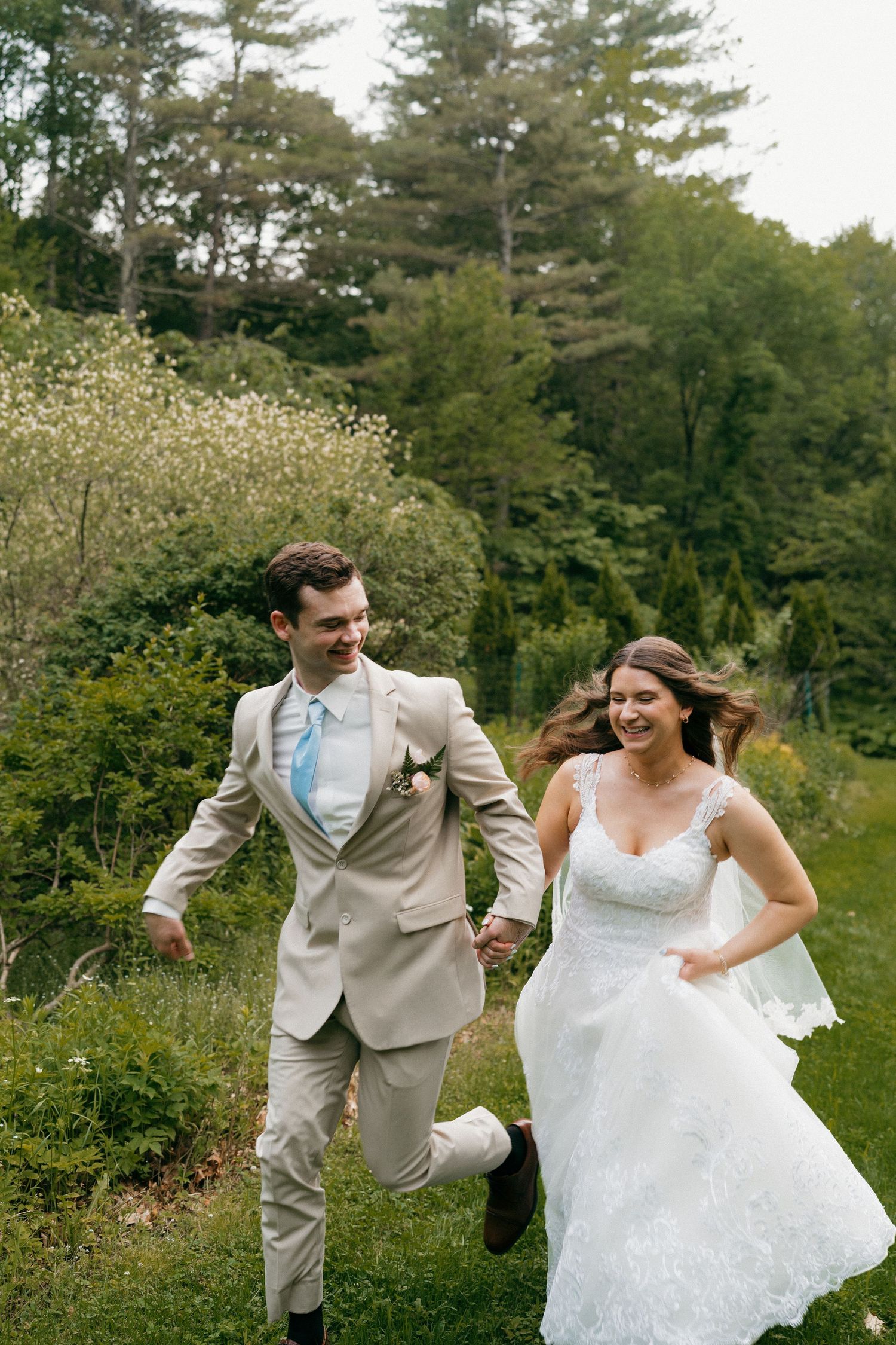 A couple in wedding attire runs playfully through a garden setting with tall pine trees in the background.