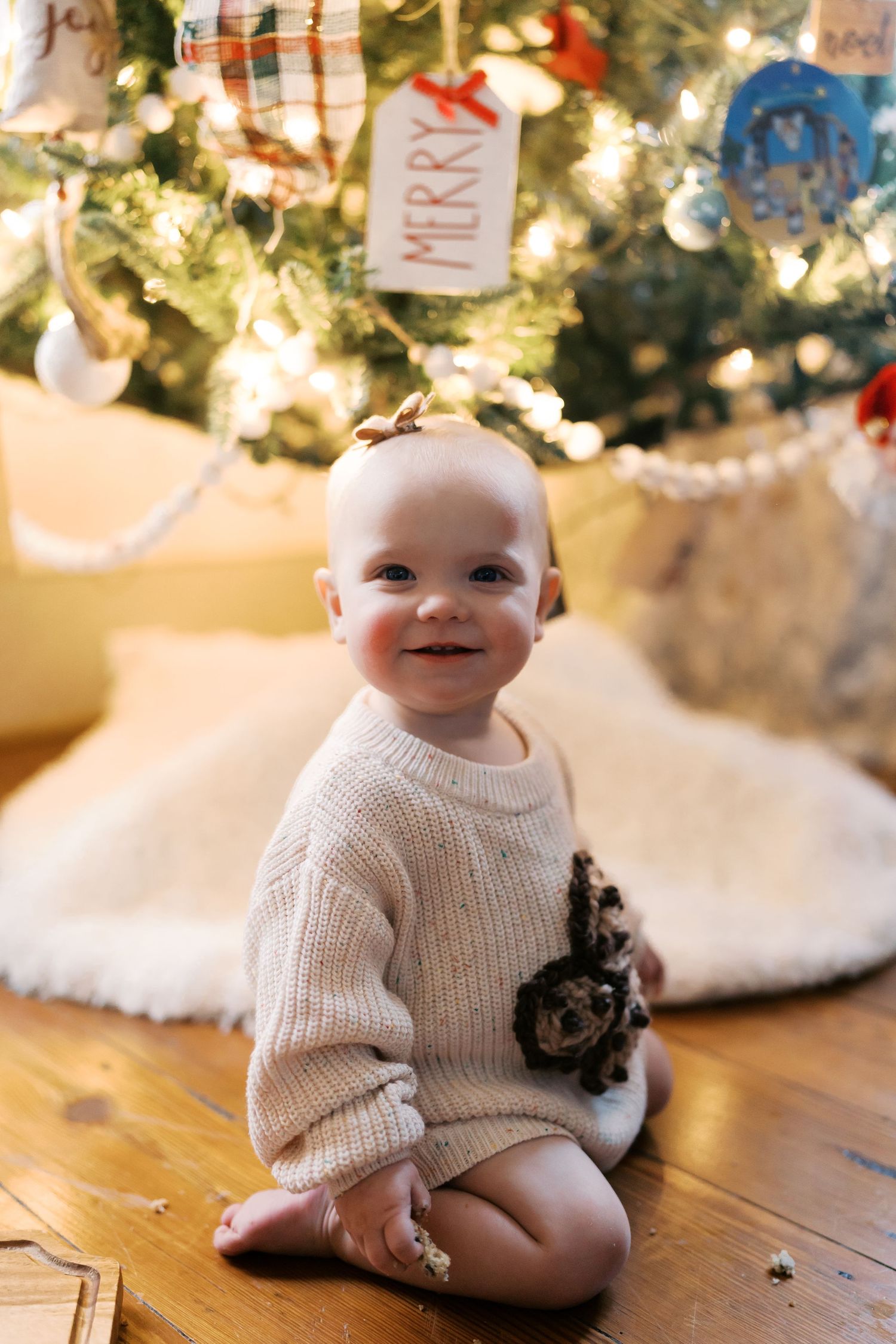 Baby in white sweater sits by illuminated Christmas tree with decorative ornaments.