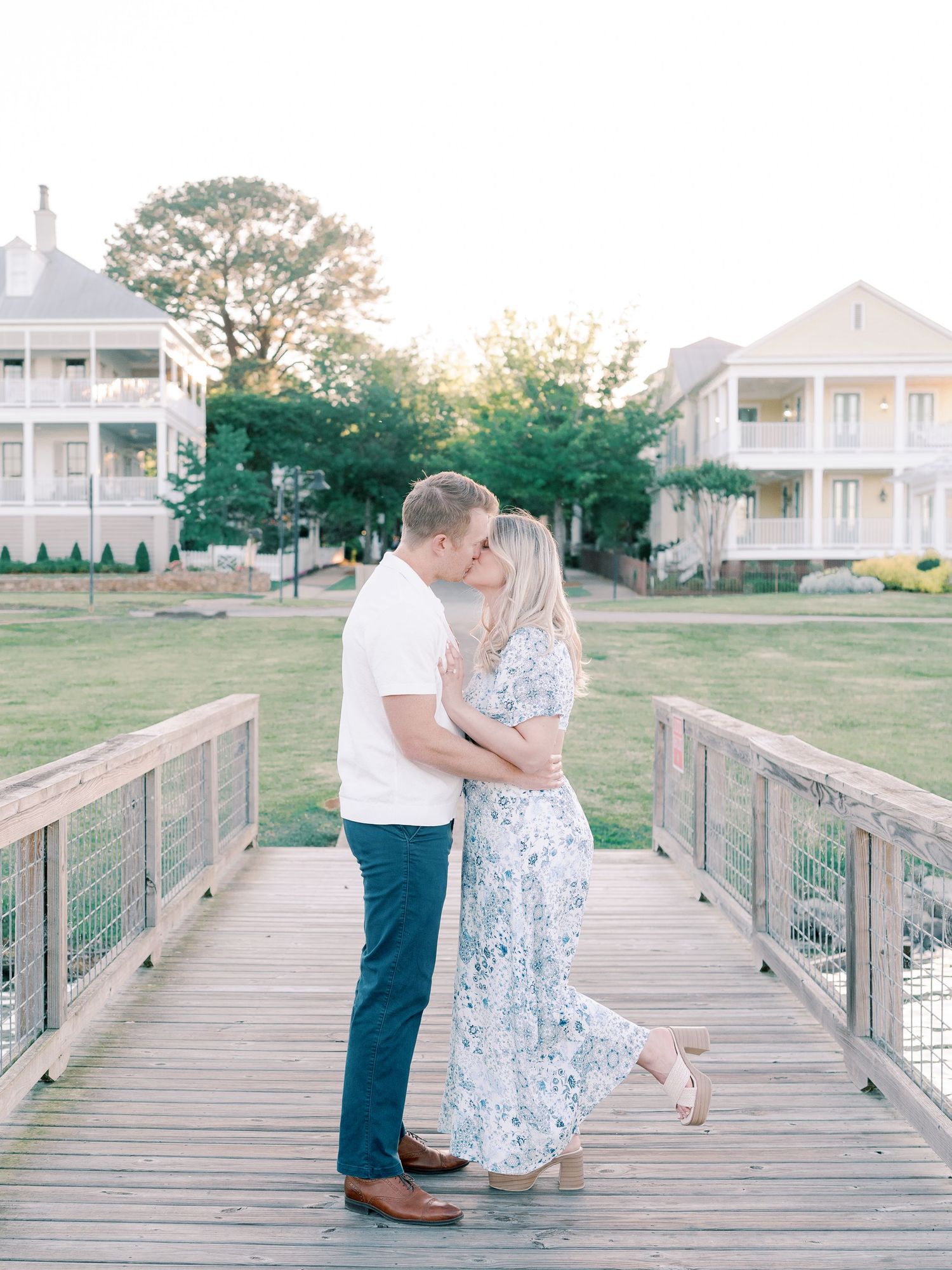 Couple poses together on wooden boardwalk with beach houses visible in distance.