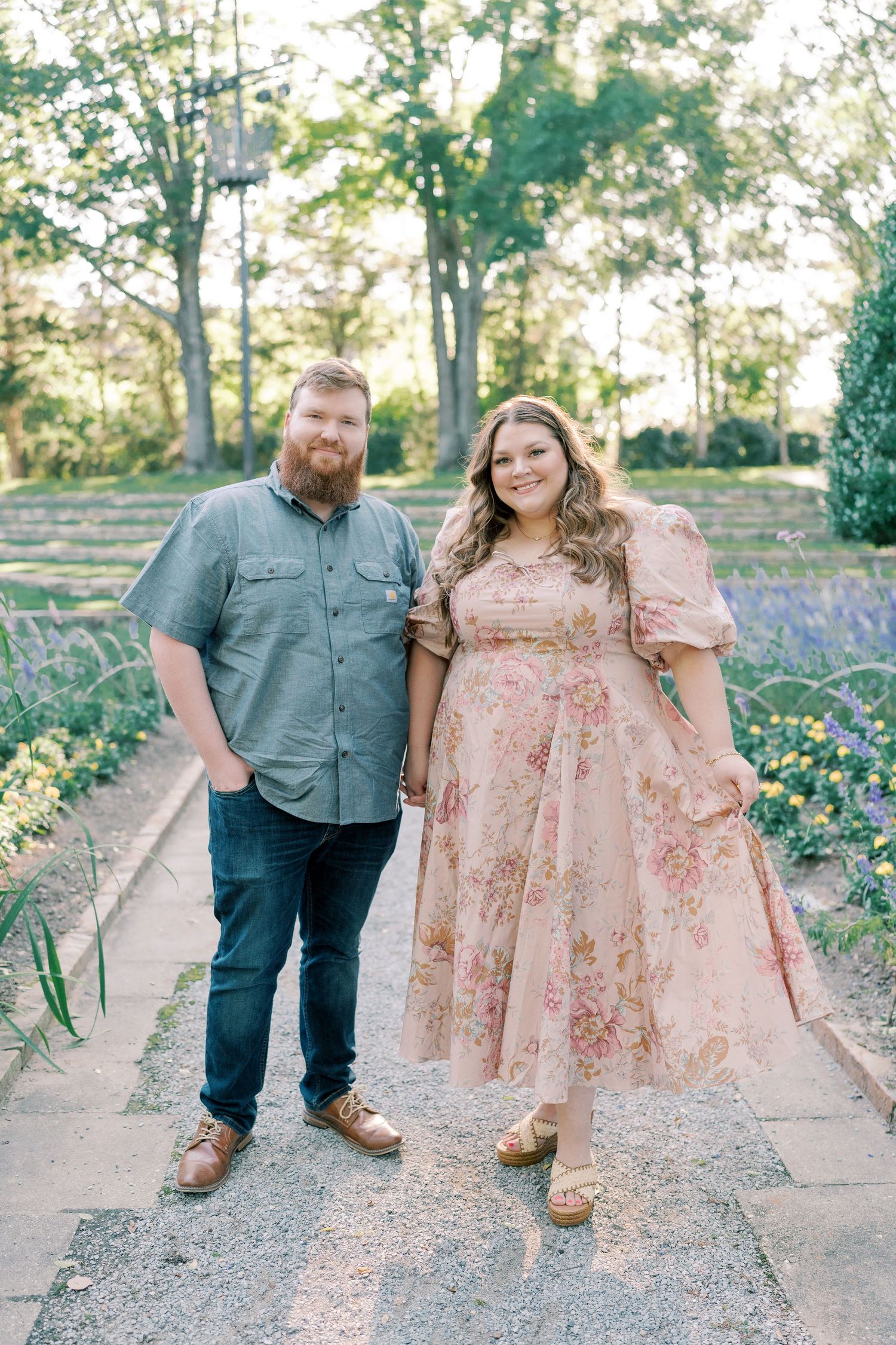 A couple posing together on a gravel path with blooming flowers and trees in the background.