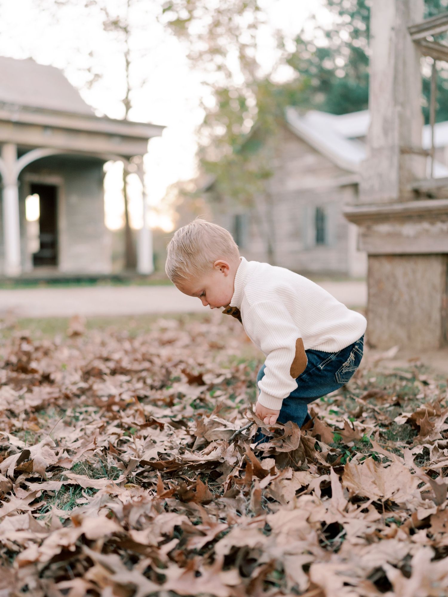 A young child bends down to play in a thick blanket of fallen autumn leaves.