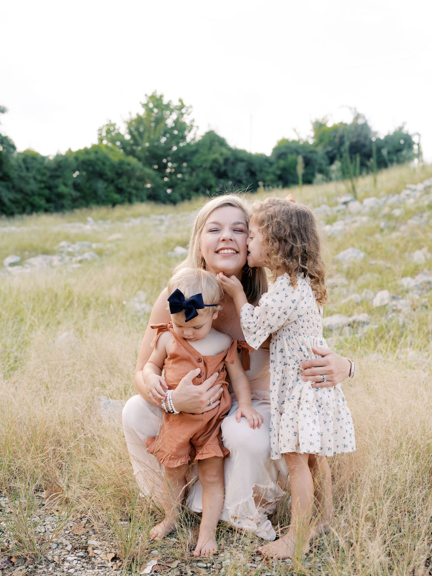 Family enjoying a candid moment together in a sunny field with tall grass and trees in the background.
