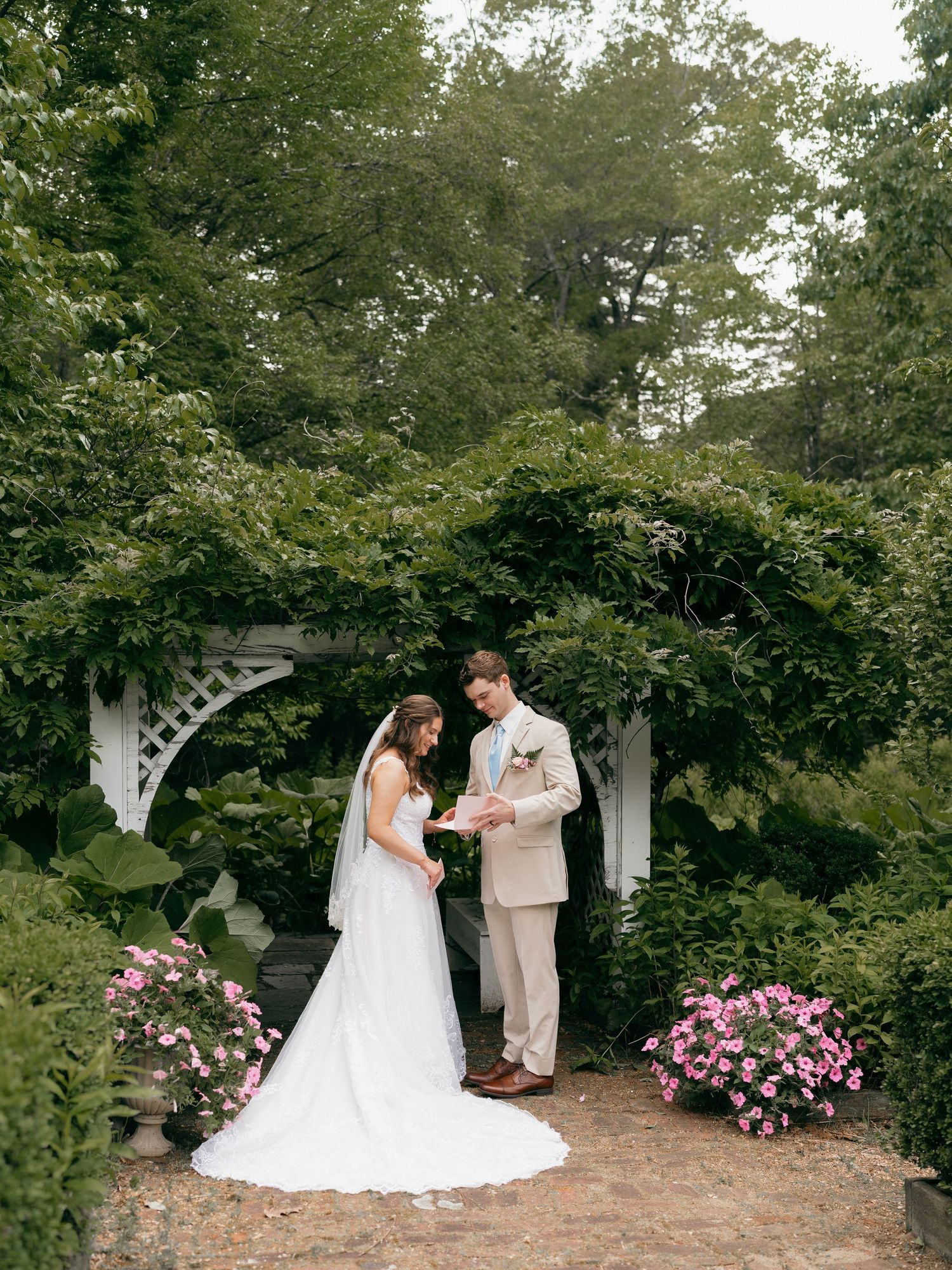 A wedding ceremony takes place under a white garden arch decorated with pink flowers.
