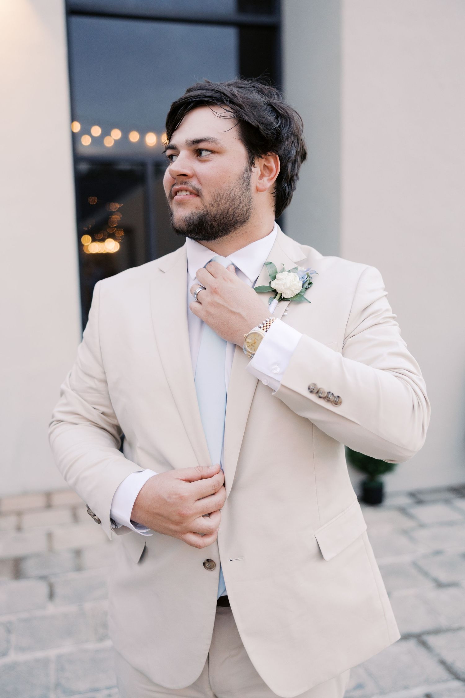 Person adjusting tie while wearing cream-colored suit jacket with floral boutonniere.