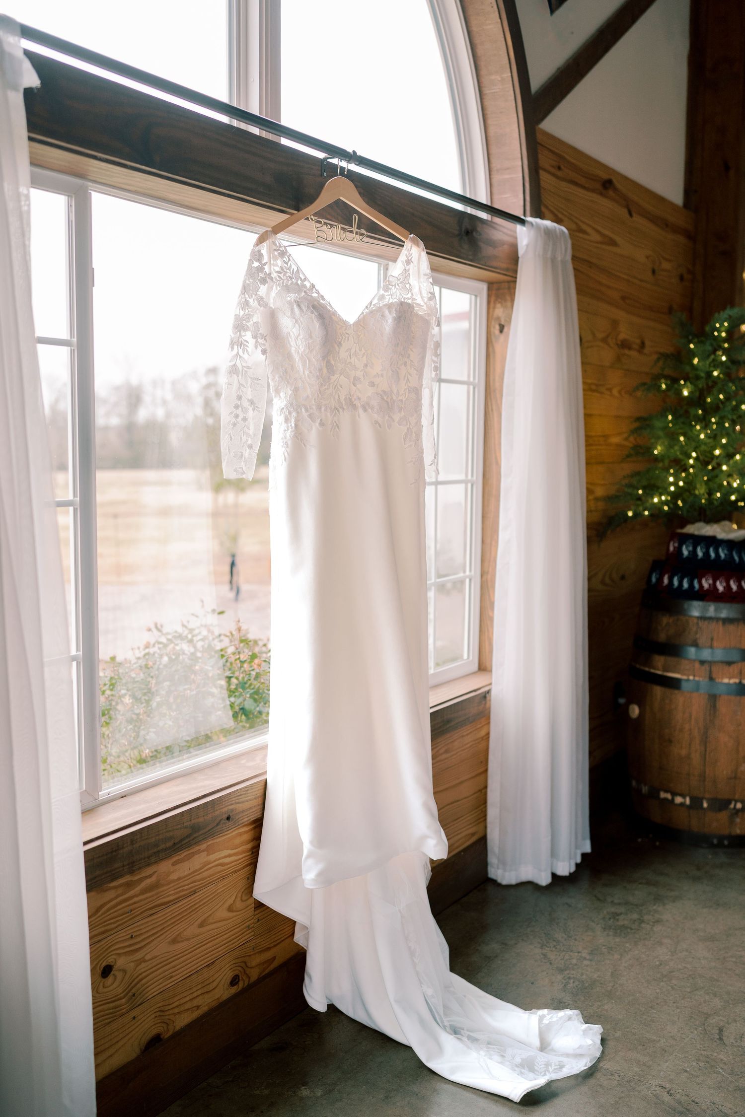 White wedding dress hangs by sunlit windows in rustic wooden room.