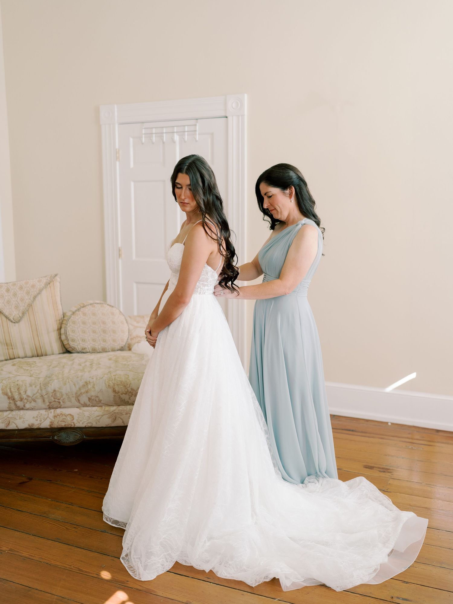 A wedding dress and bridesmaid in light blue gown prepare in an elegant white room.