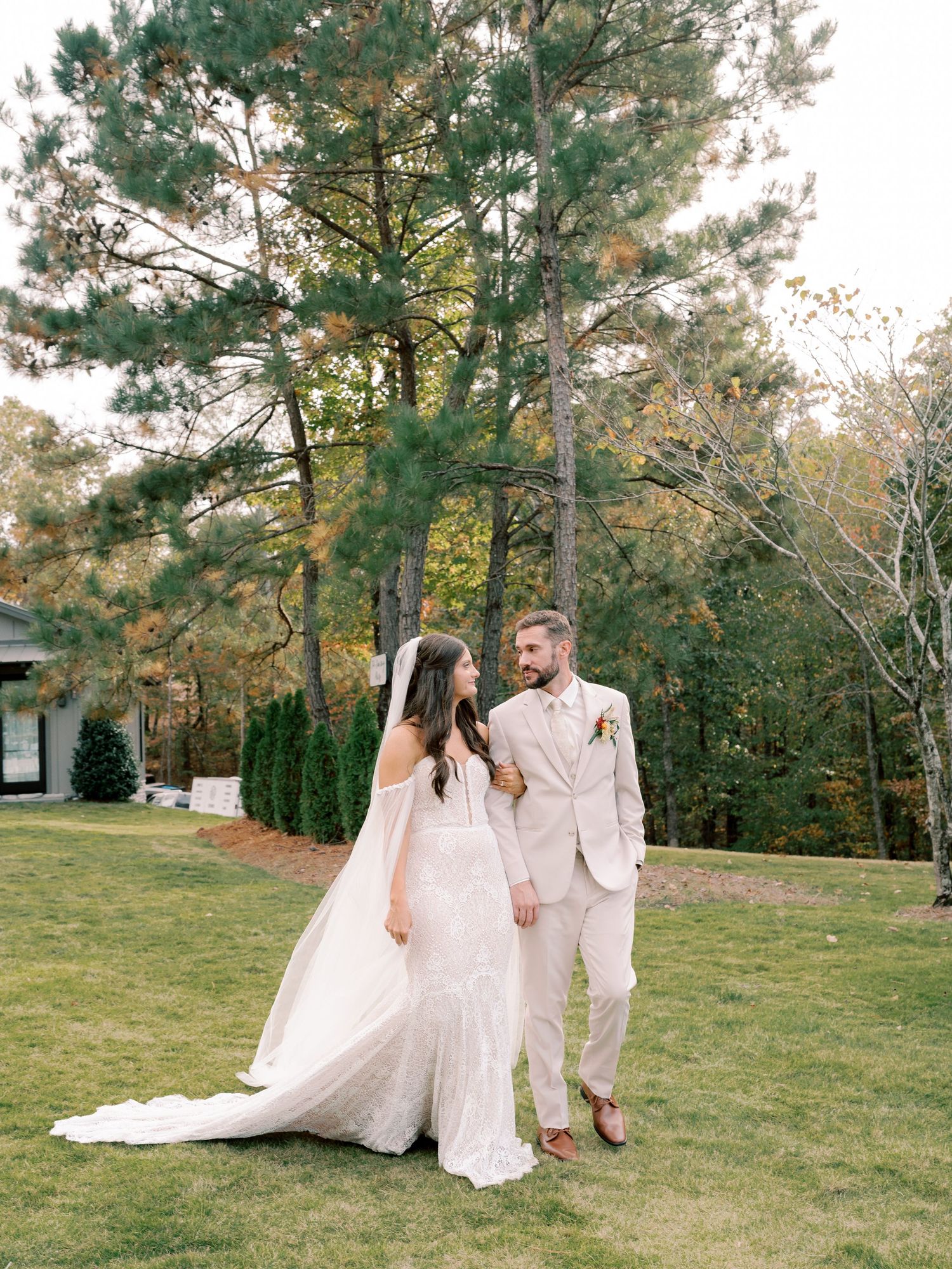 Couple in wedding attire walking hand in hand across a grassy lawn with pine trees in background.