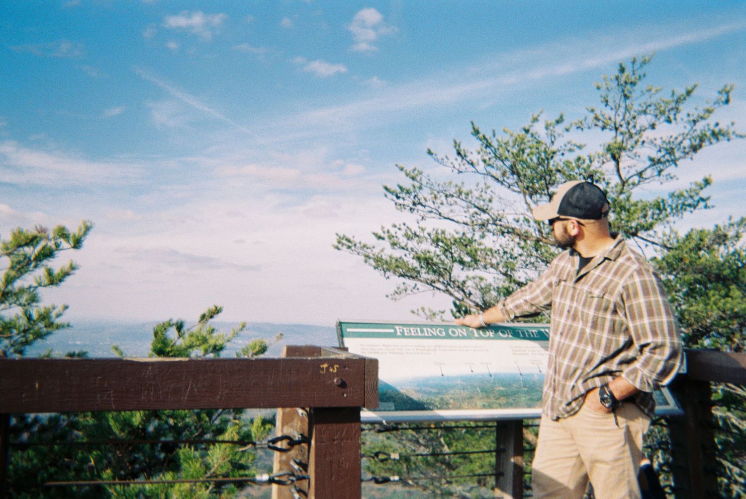 Person in plaid shirt and baseball cap looking out over scenic mountain vista.