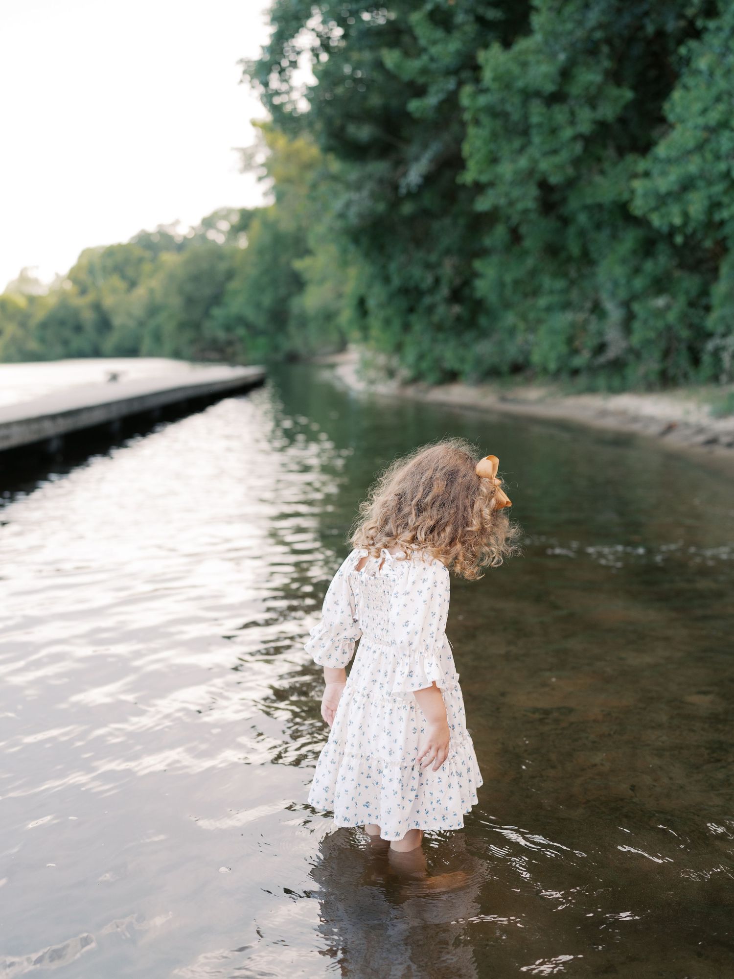 Child in white dress playing and splashing in shallow river water near a wooded shore.