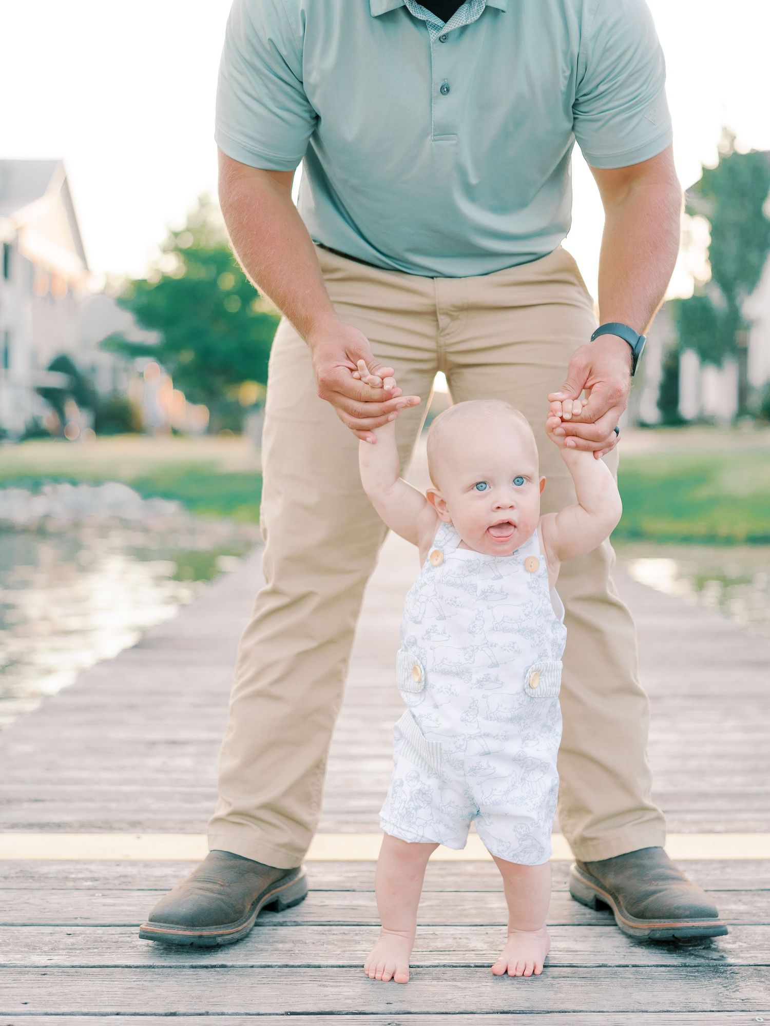 Baby in white romper learning to walk between adult legs on wooden dock.