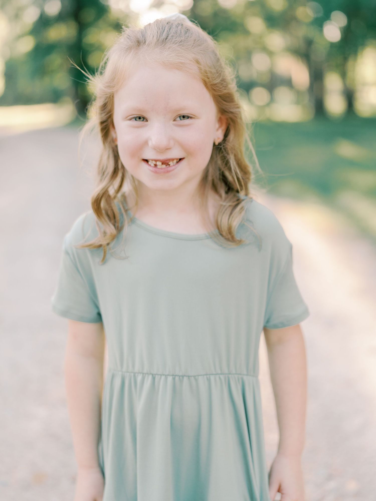 Young child in a sage green dress smiling brightly outdoors in summer sunlight.