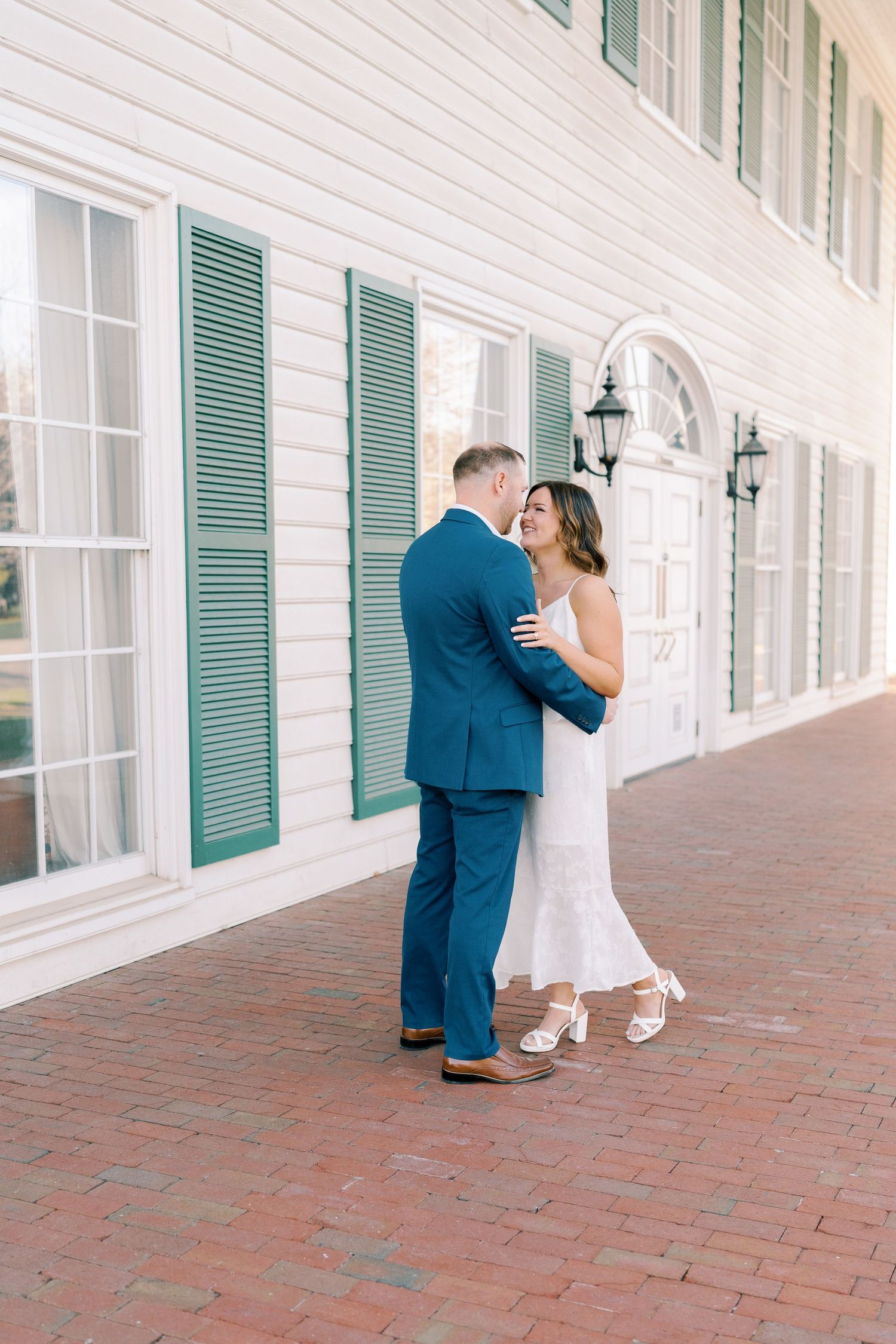 Couple embraces on brick porch with white walls and green shutters in elegant setting.