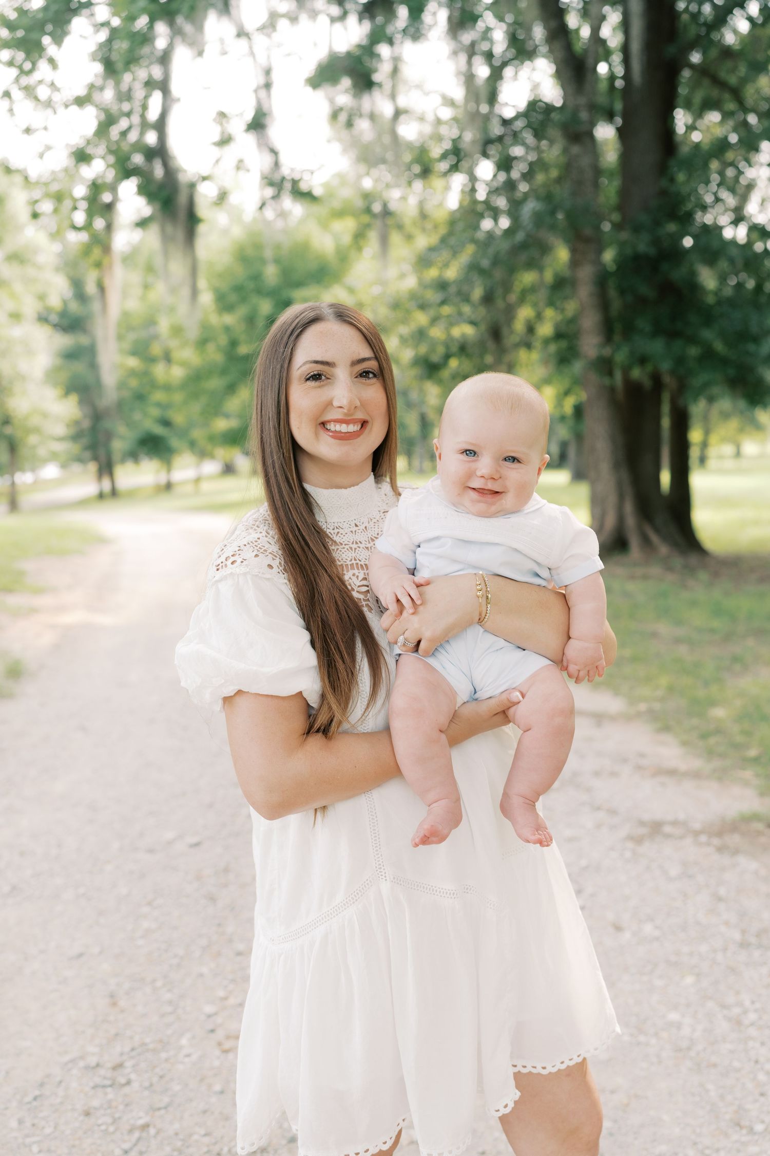 A woman in a white lace dress holds a smiling baby on a park trail.