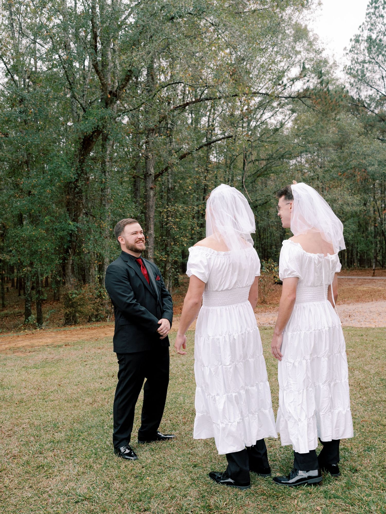 Wedding party in white dresses and formal attire poses in outdoor woodland setting.