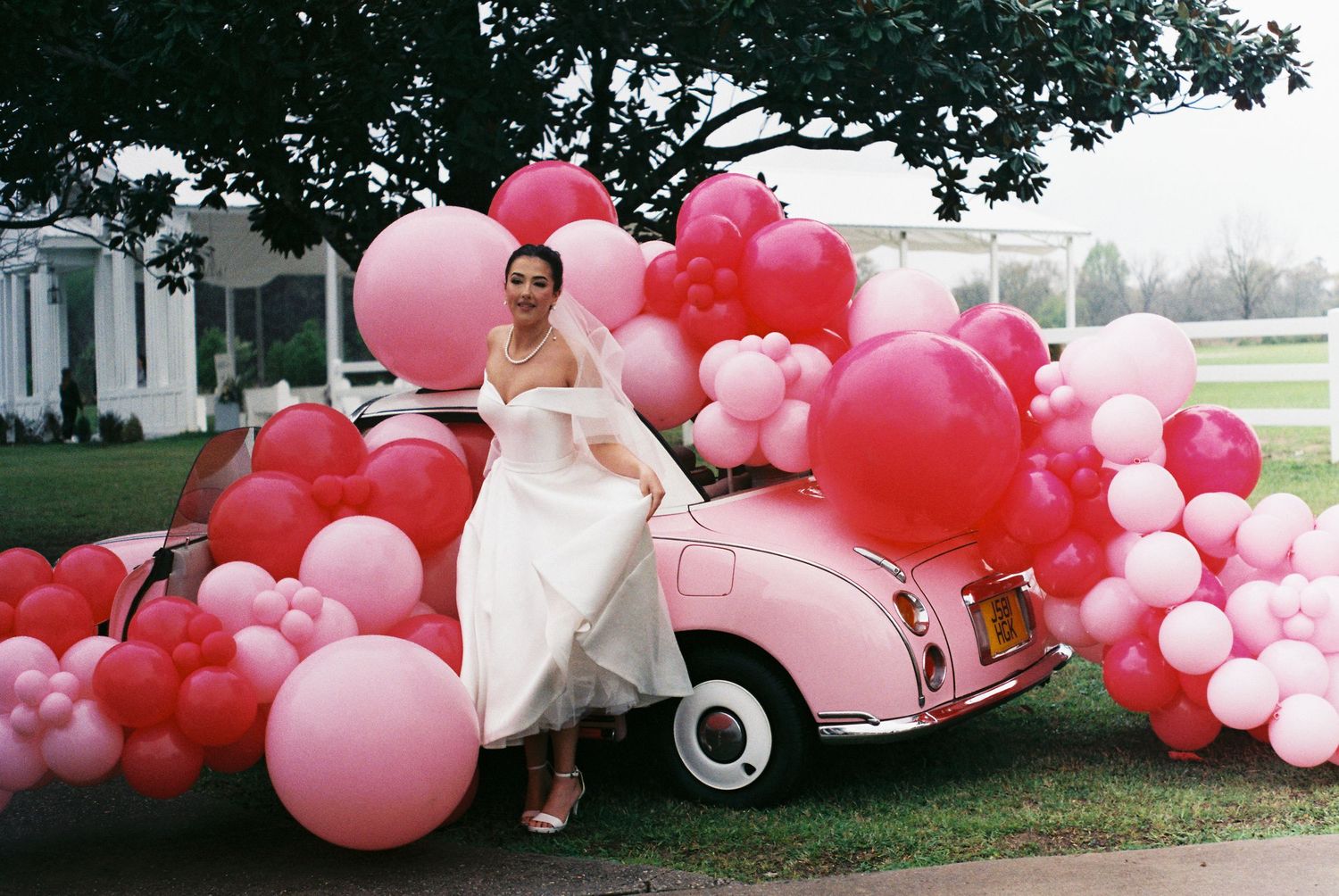 Pink and red balloon arch decorating a vintage pink car at an outdoor event.