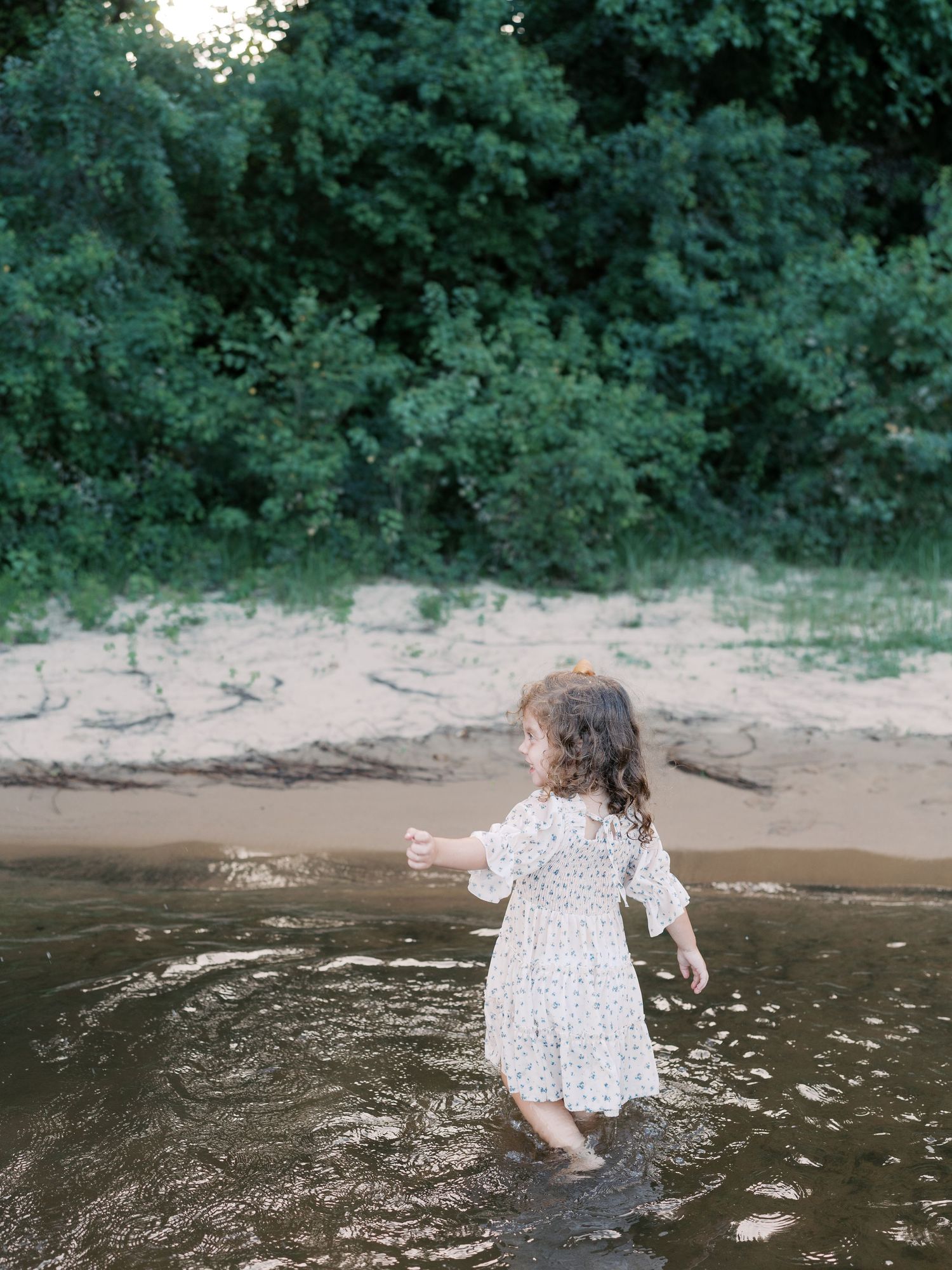 Child in white dress exploring shallow river water near a sandy beach.