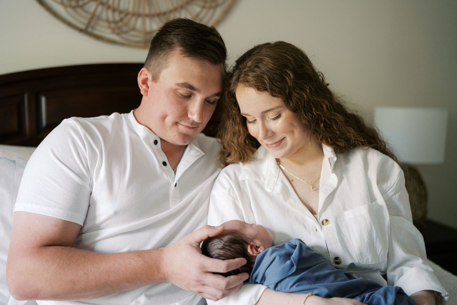 Couple relaxing together on a white bed with their baby in a cozy bedroom setting.