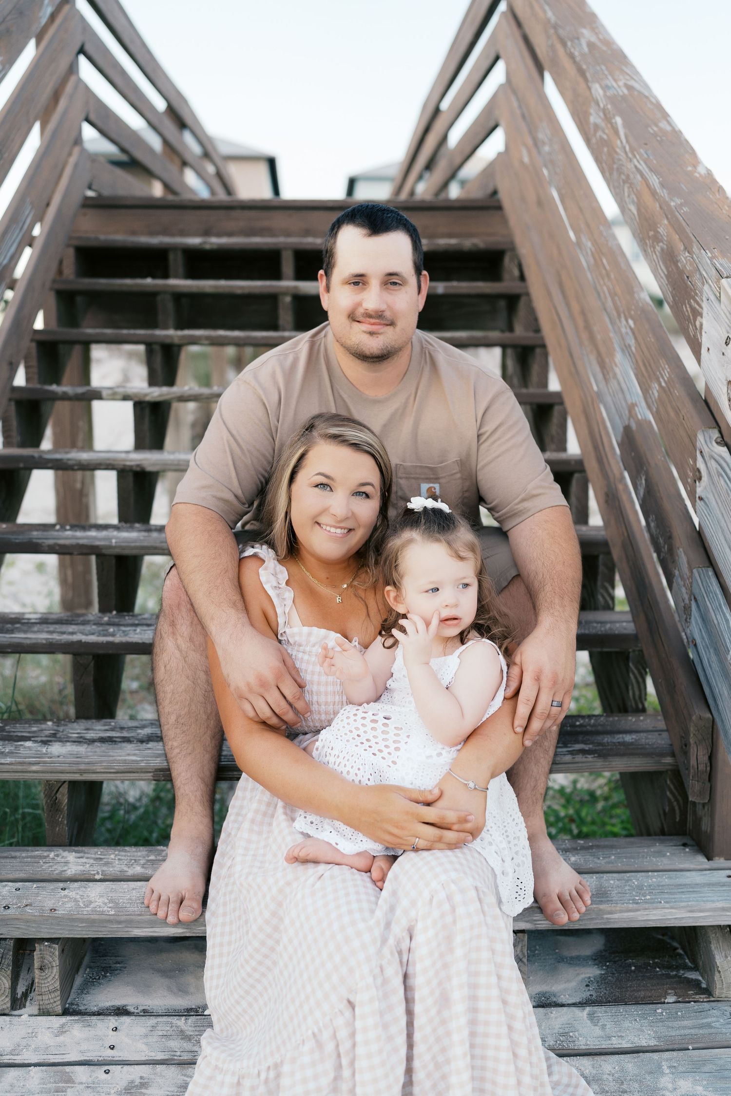 A family poses together on wooden stairs in coordinated white and tan outfits.