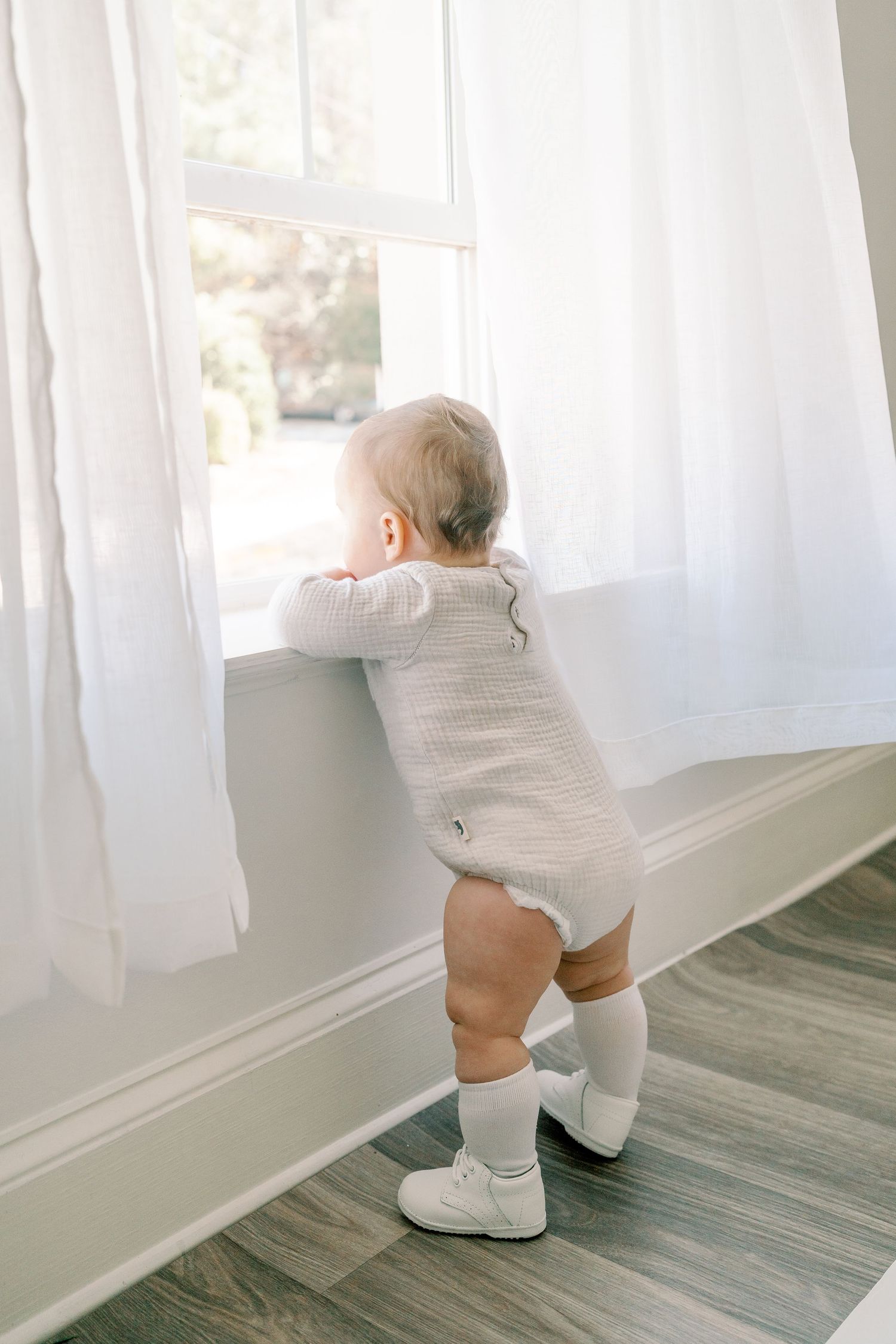 Baby in white outfit exploring sheer curtains by window with natural light.