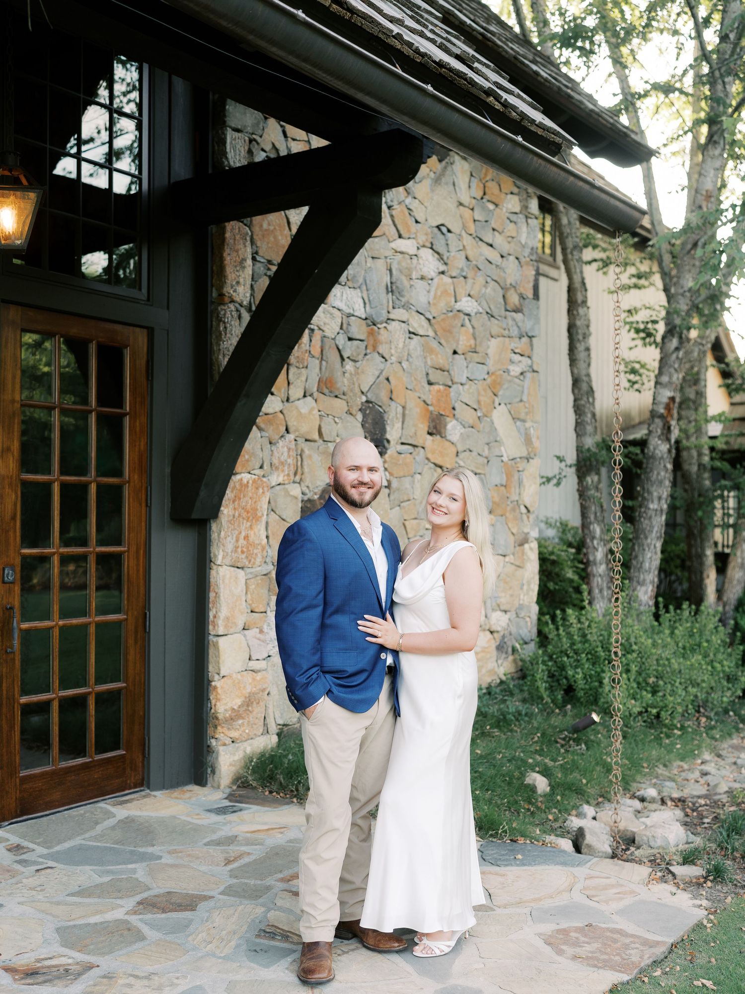 Couple poses together against rustic stone building with wooden windows.