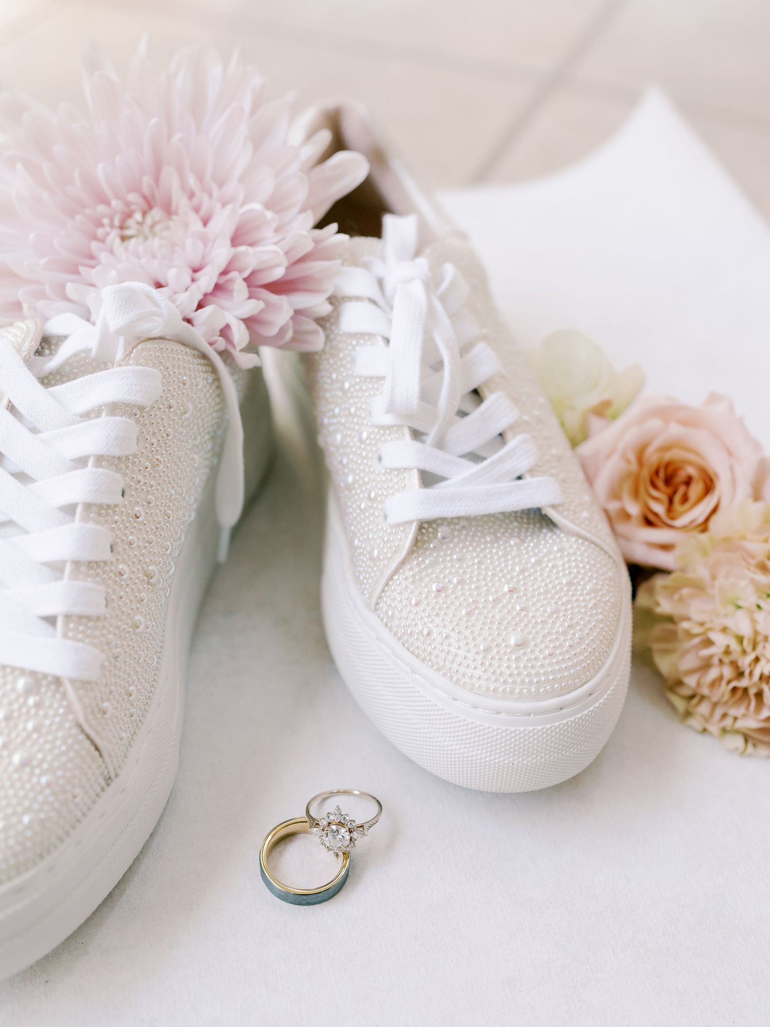White bridal sneakers with rhinestones next to wedding rings and pink flowers.