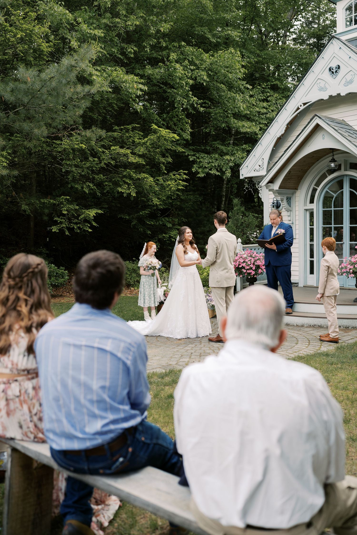 Wedding ceremony in progress outside a white chapel with guests seated on rustic benches.